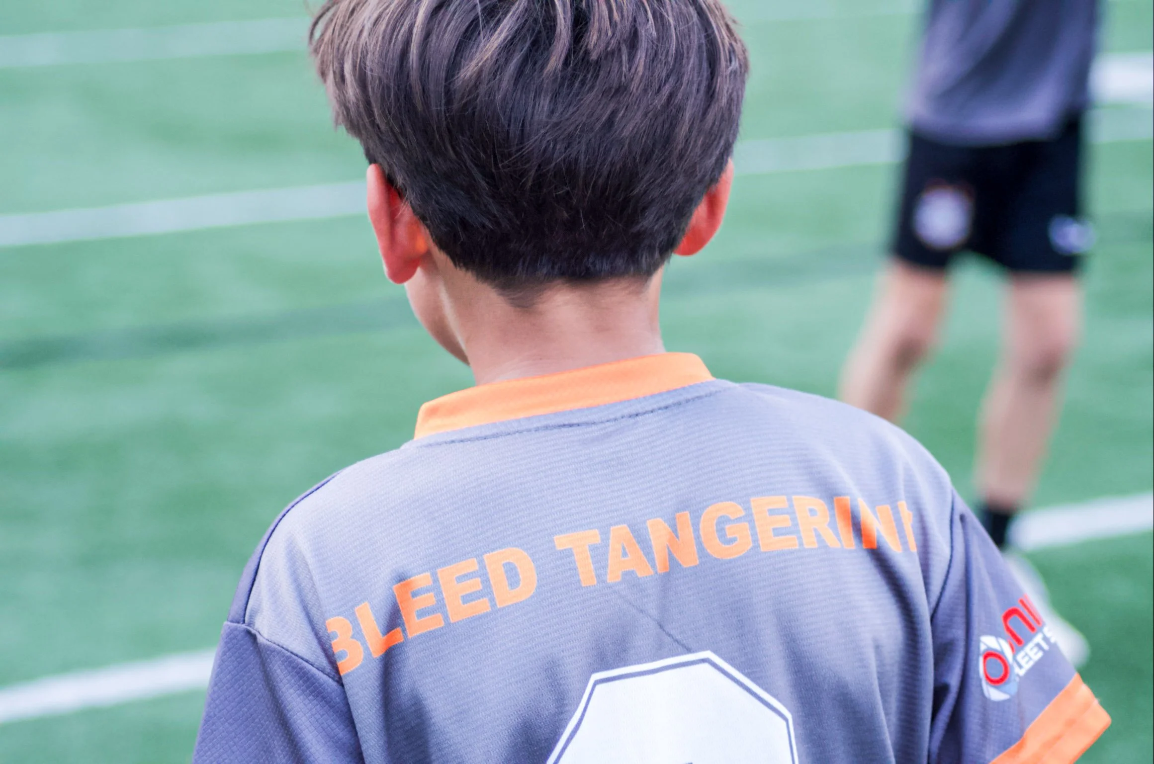The back of a young soccer player wearing a gray jersey with orange lettering that says 'Bleed Tangerine,' with a blurred soccer field and other players in the background.