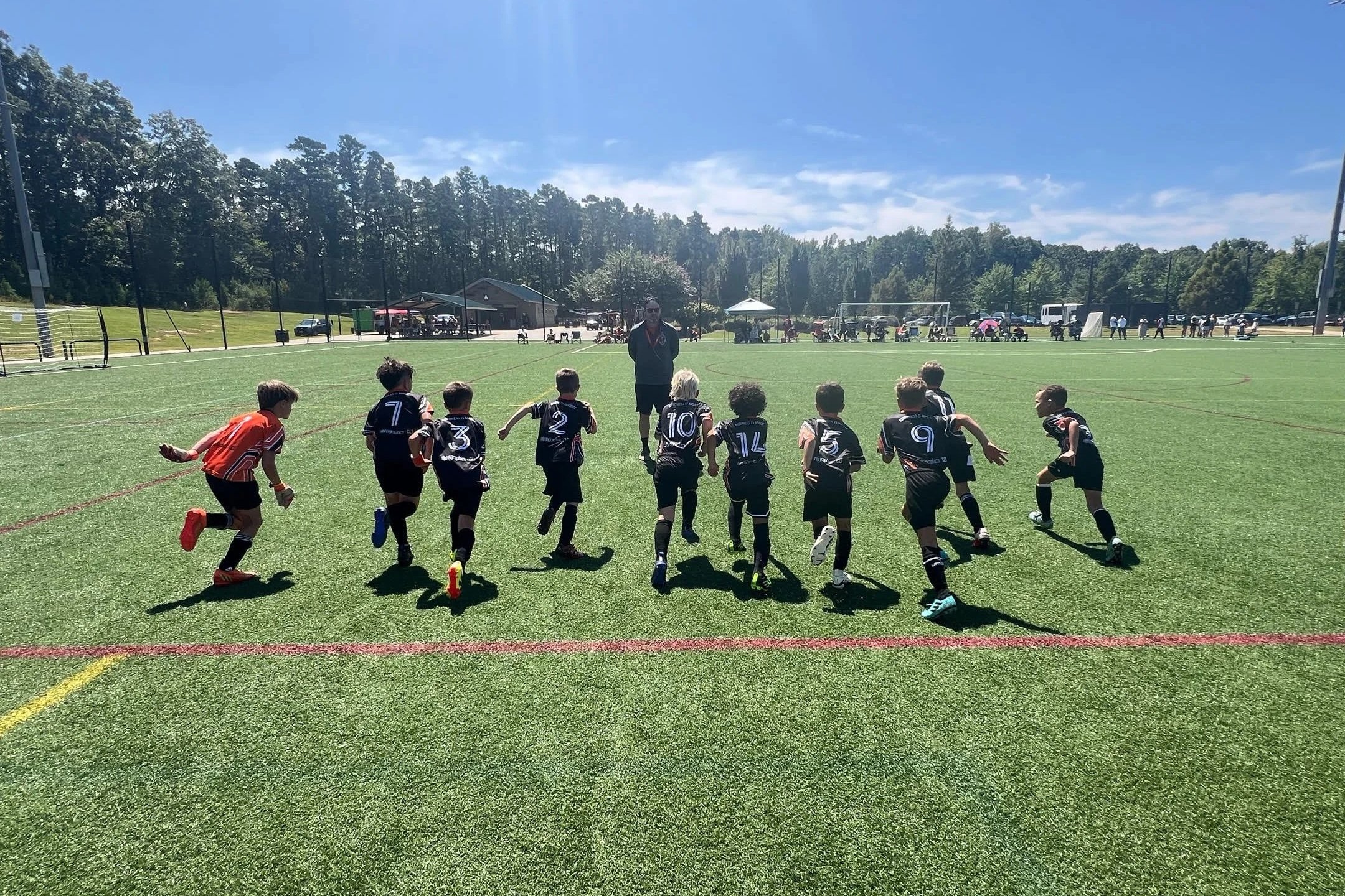 Children playing soccer on a sunny day, with a coach standing nearby on a grass field.