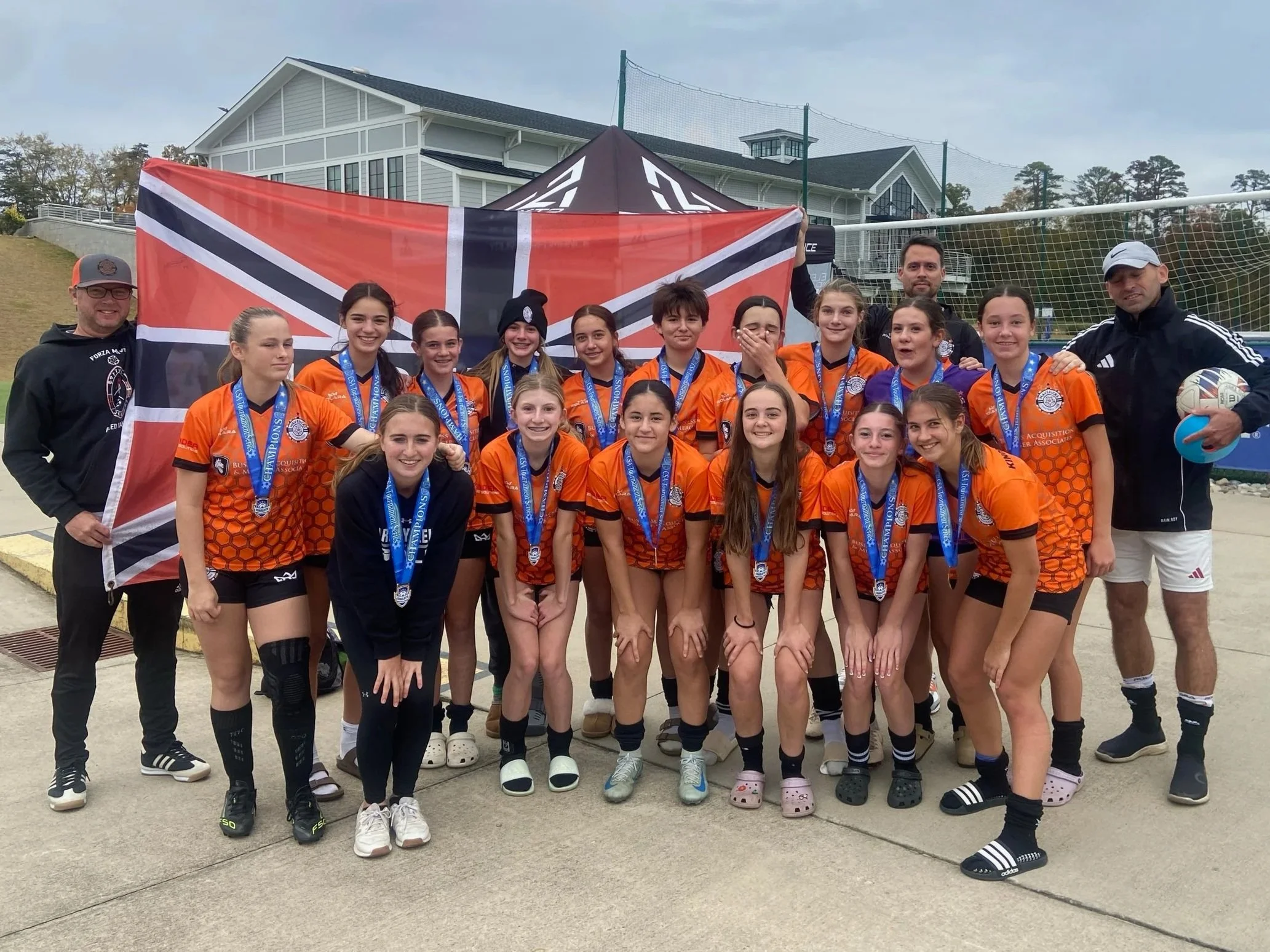 A girls' soccer team with coaches celebrating after a game or tournament, wearing orange jerseys and medals, holding a flag and a volleyball, outside near a sports field or complex.
