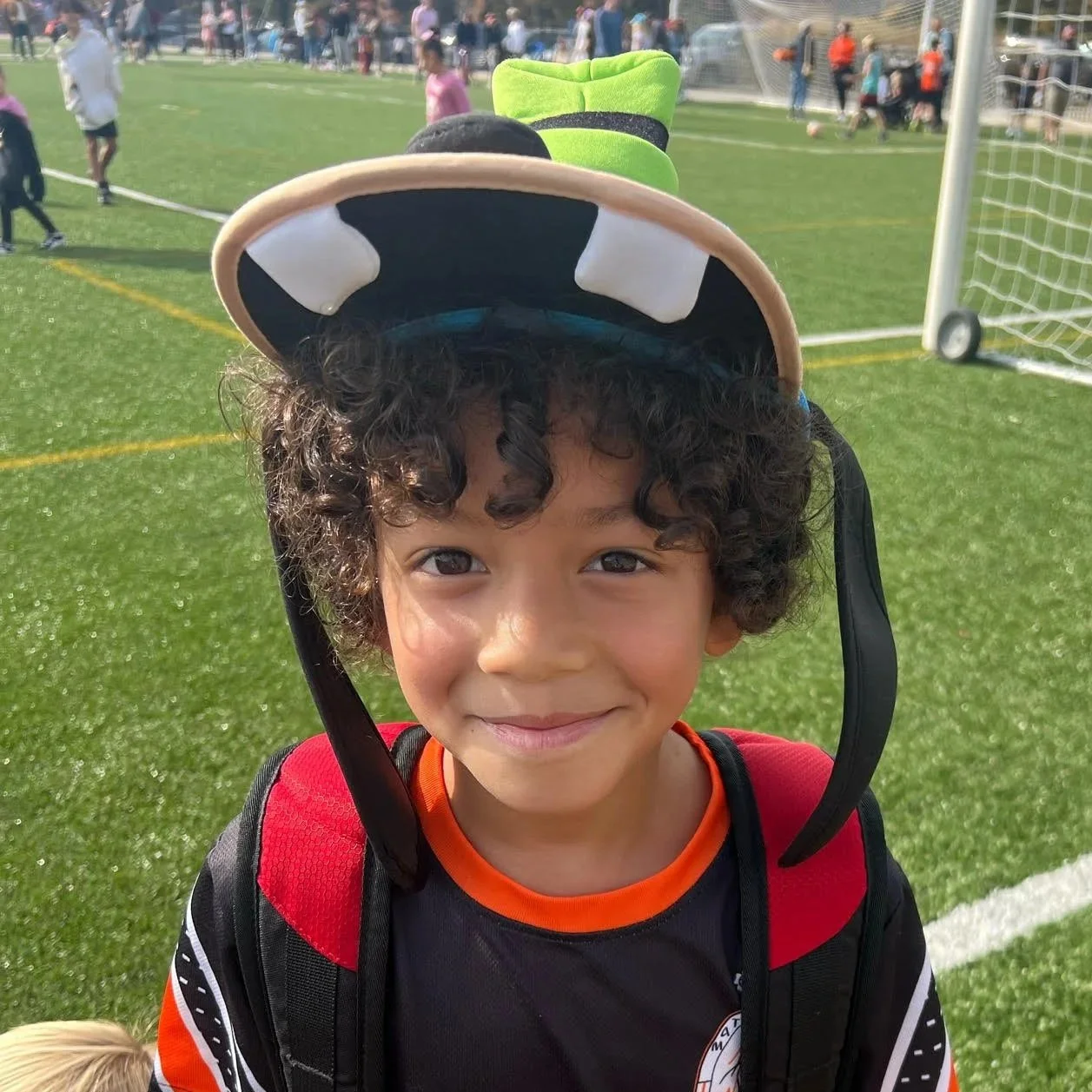 A young boy with curly hair and a red and black sports jersey smiling at the camera, wearing a cap with a cow face design. He is standing on a grassy sports field with other children and adults in the background.