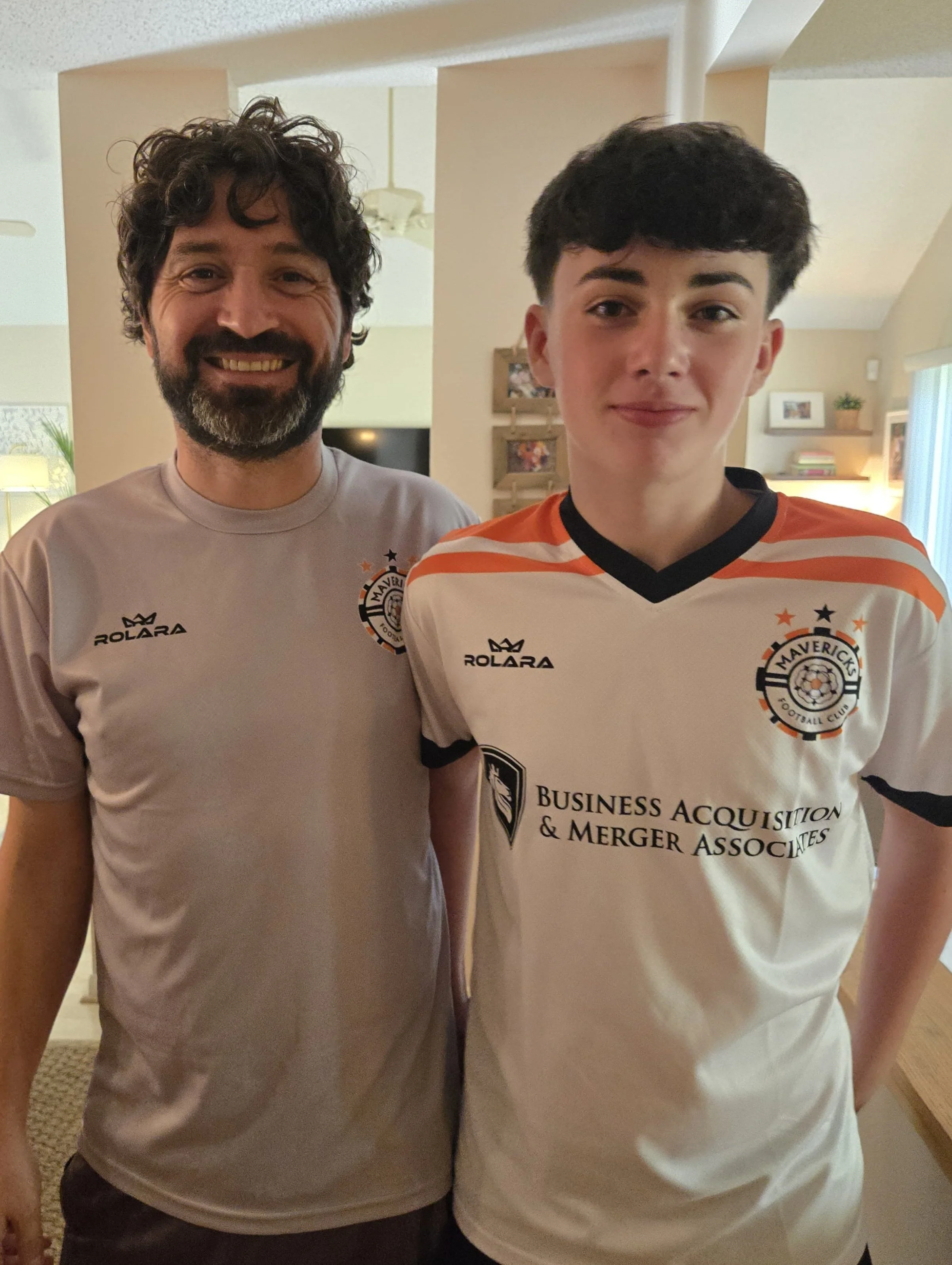 Two people standing indoors, wearing Maverick Football Club jerseys, smiling at the camera.