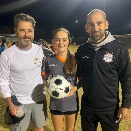 A young female soccer player holding a soccer ball, standing between two adult men on a soccer field at night.