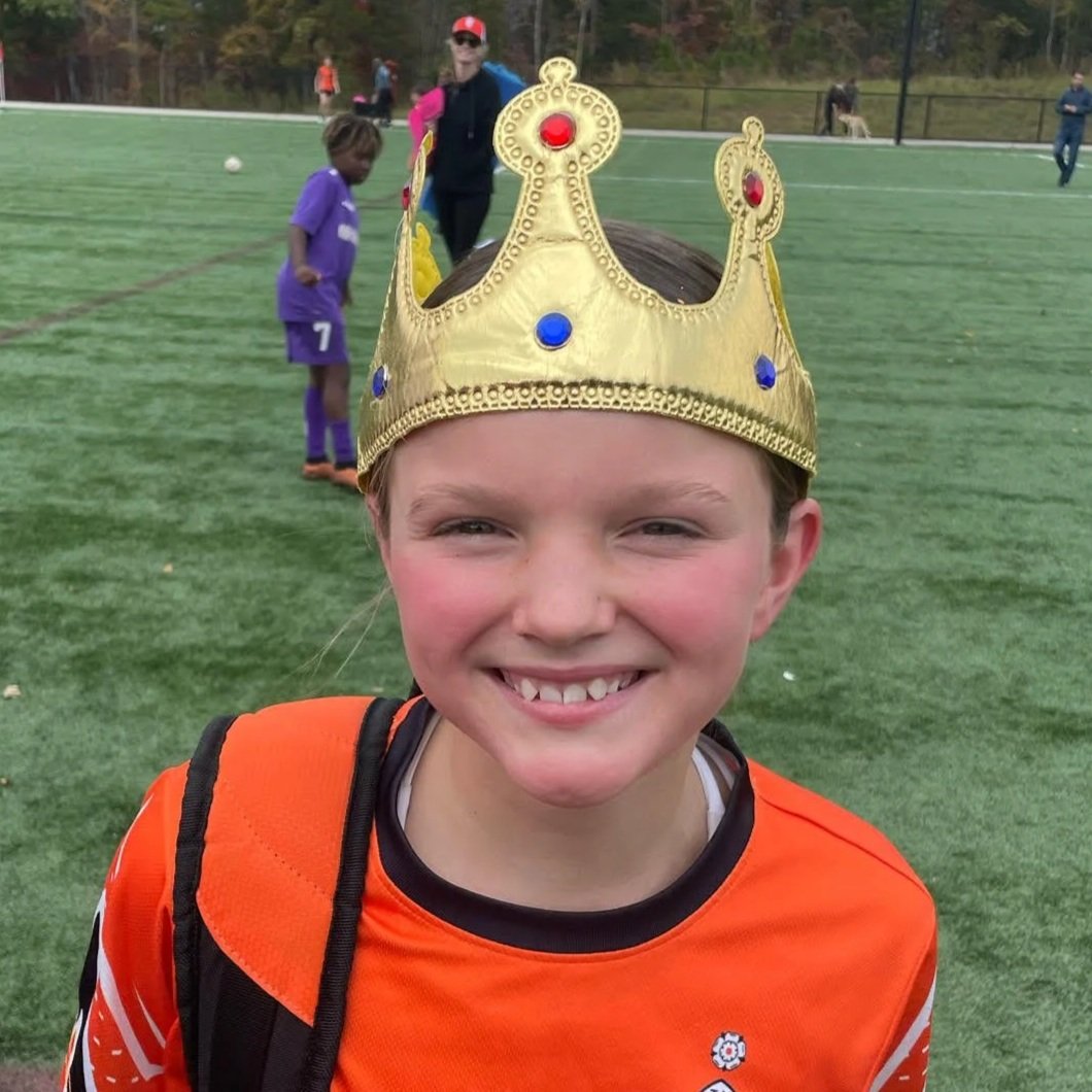A young boy wearing an orange sports jersey and a gold crown, smiling on a soccer field.