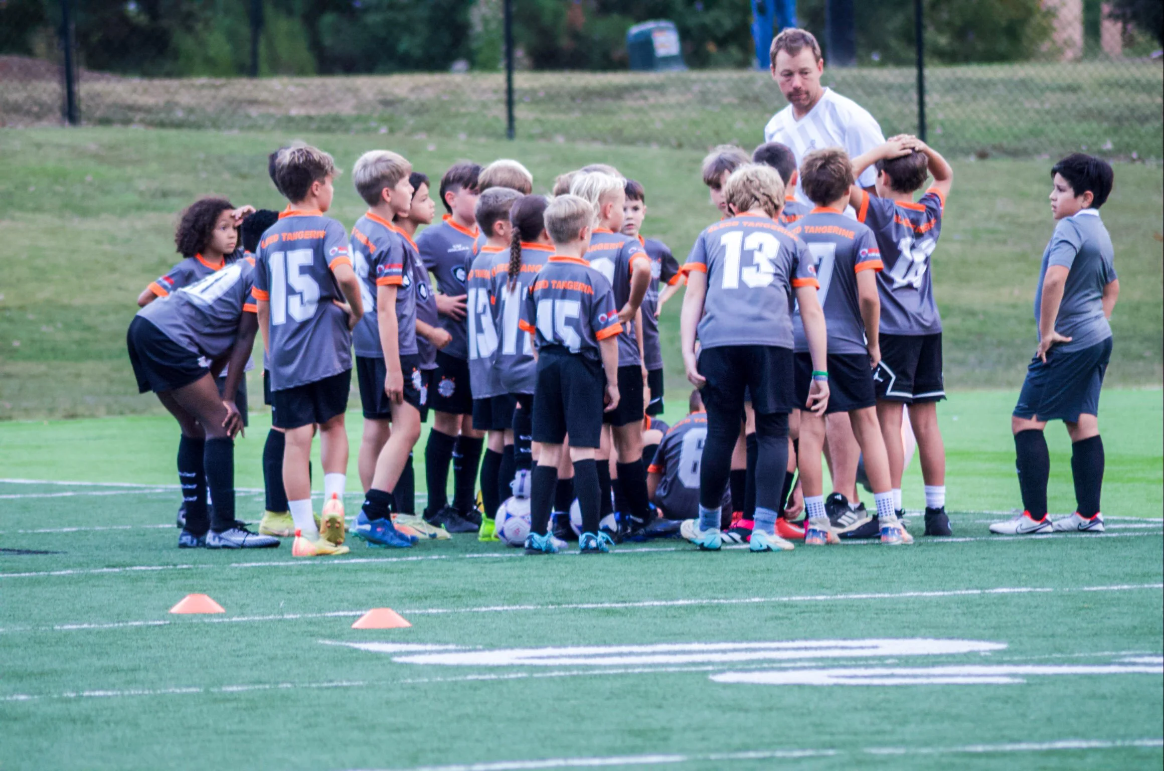 A youth soccer team with their coach on a field during practice or a game, with players wearing gray and black uniforms with orange accents, standing around a player on the ground, while one coach or adult stands nearby.