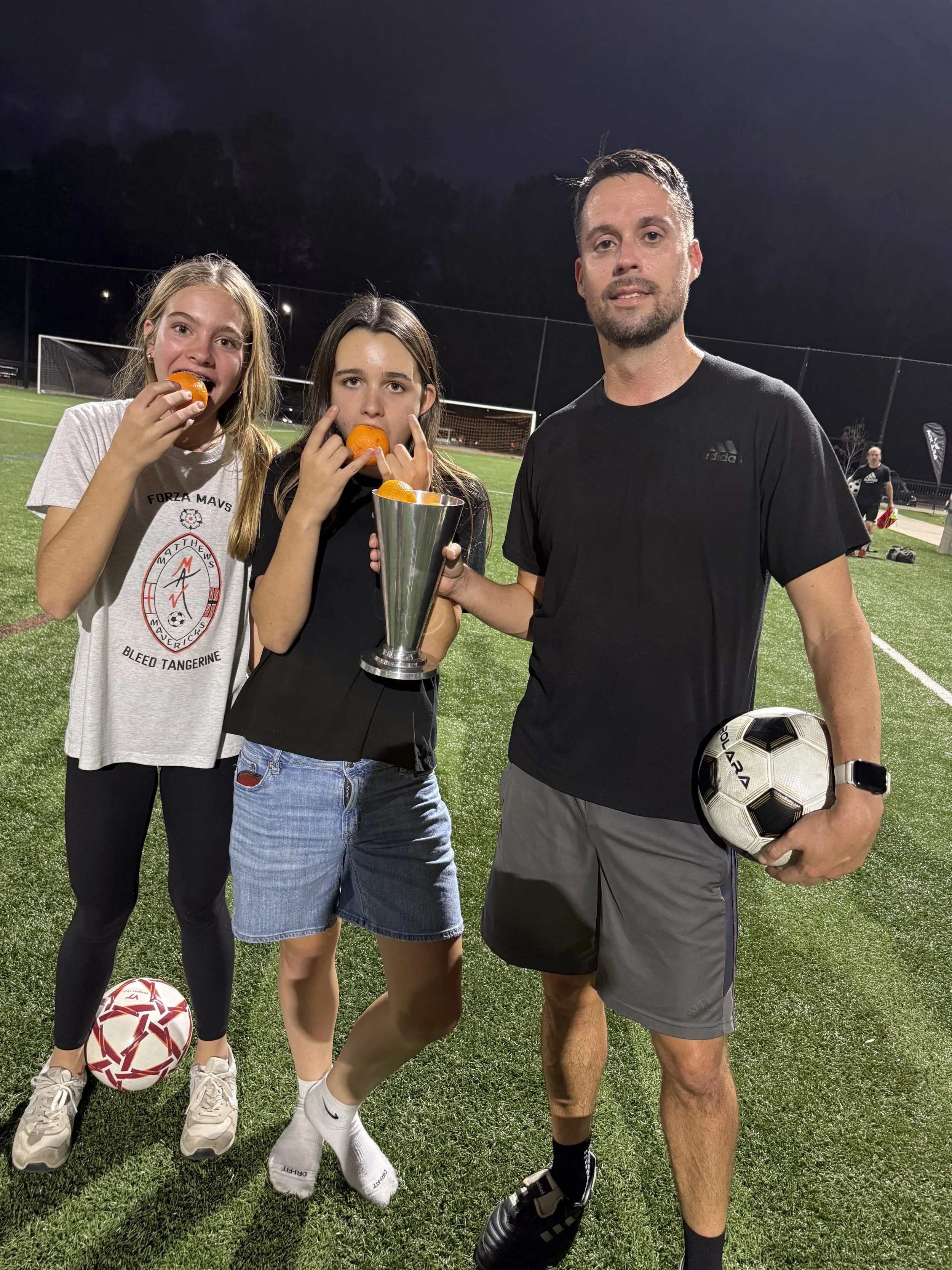Three people standing on a soccer field at night, holding a trophy and soccer balls, celebrating a victory.