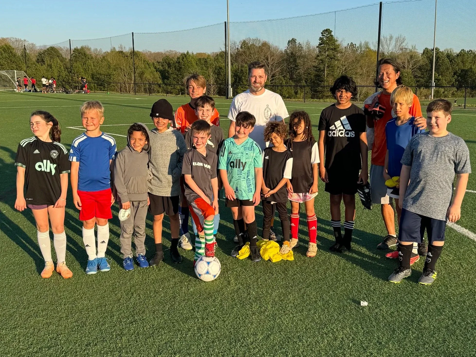 Group of children and two adults standing on a soccer field, with some children wearing sports jerseys, and one child balancing a soccer ball on their foot.