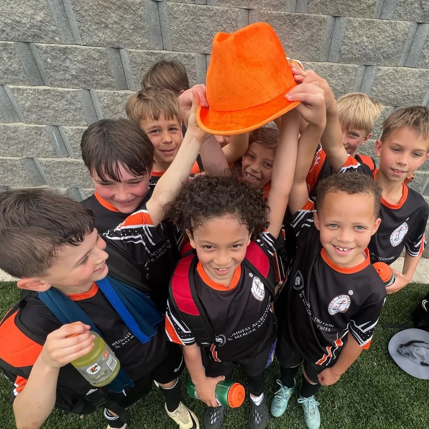 Group of young boys in black and orange sports jerseys celebrating, holding an orange hat in the air, standing outside against a stone wall.
