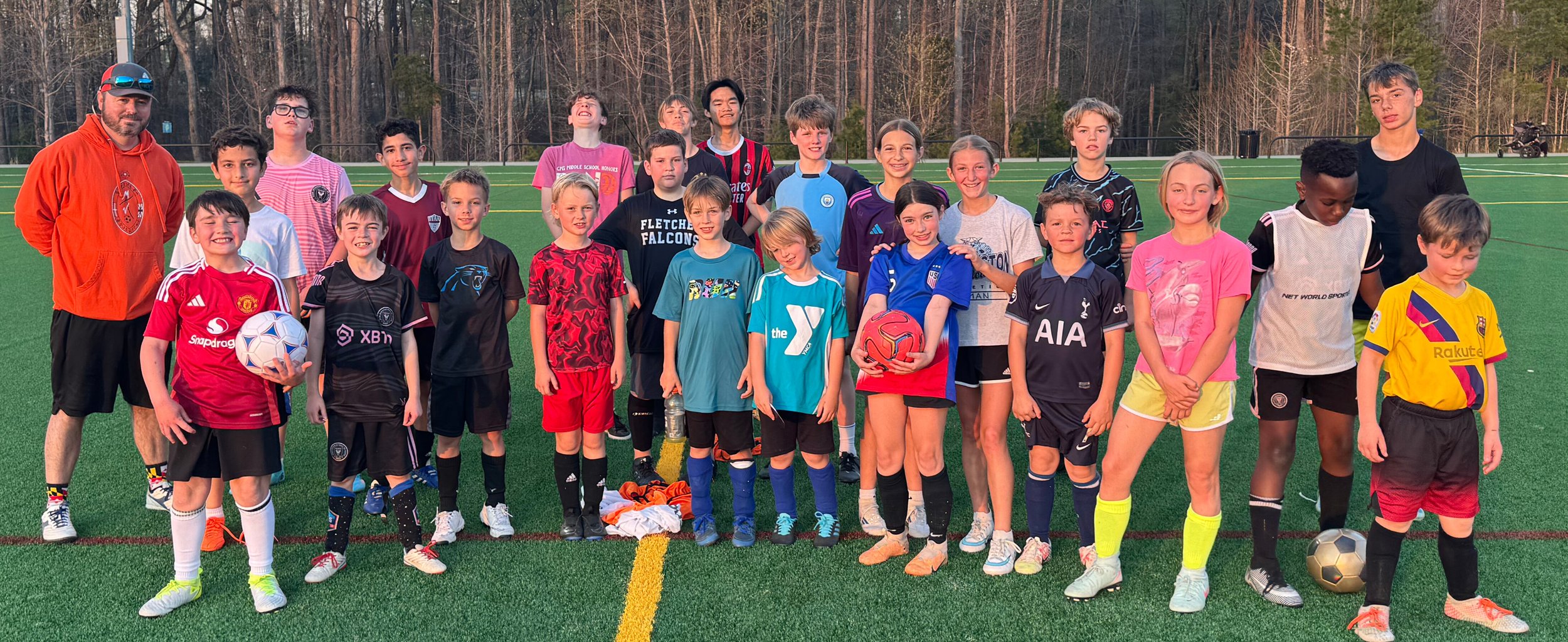 A group of children and two adults on a green synthetic soccer field with a backdrop of leafless trees, posing for a team photo. Some children are holding soccer balls, and they are wearing various colorful sports jerseys and shorts.
