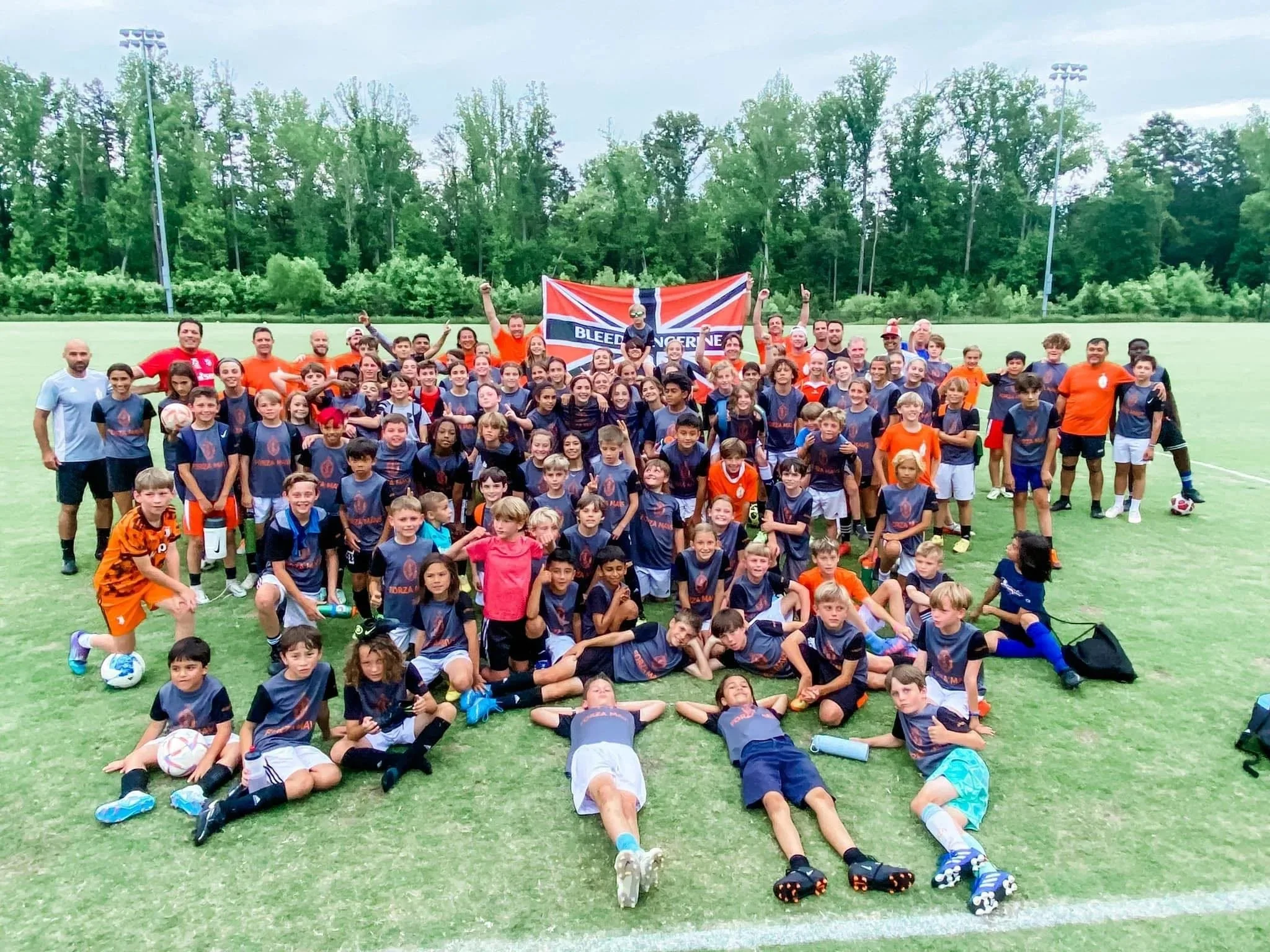 Large group of children and adults on a soccer field, posing for a team photo. Many are wearing matching jerseys, and some are holding soccer balls. There are trees and floodlights in the background.