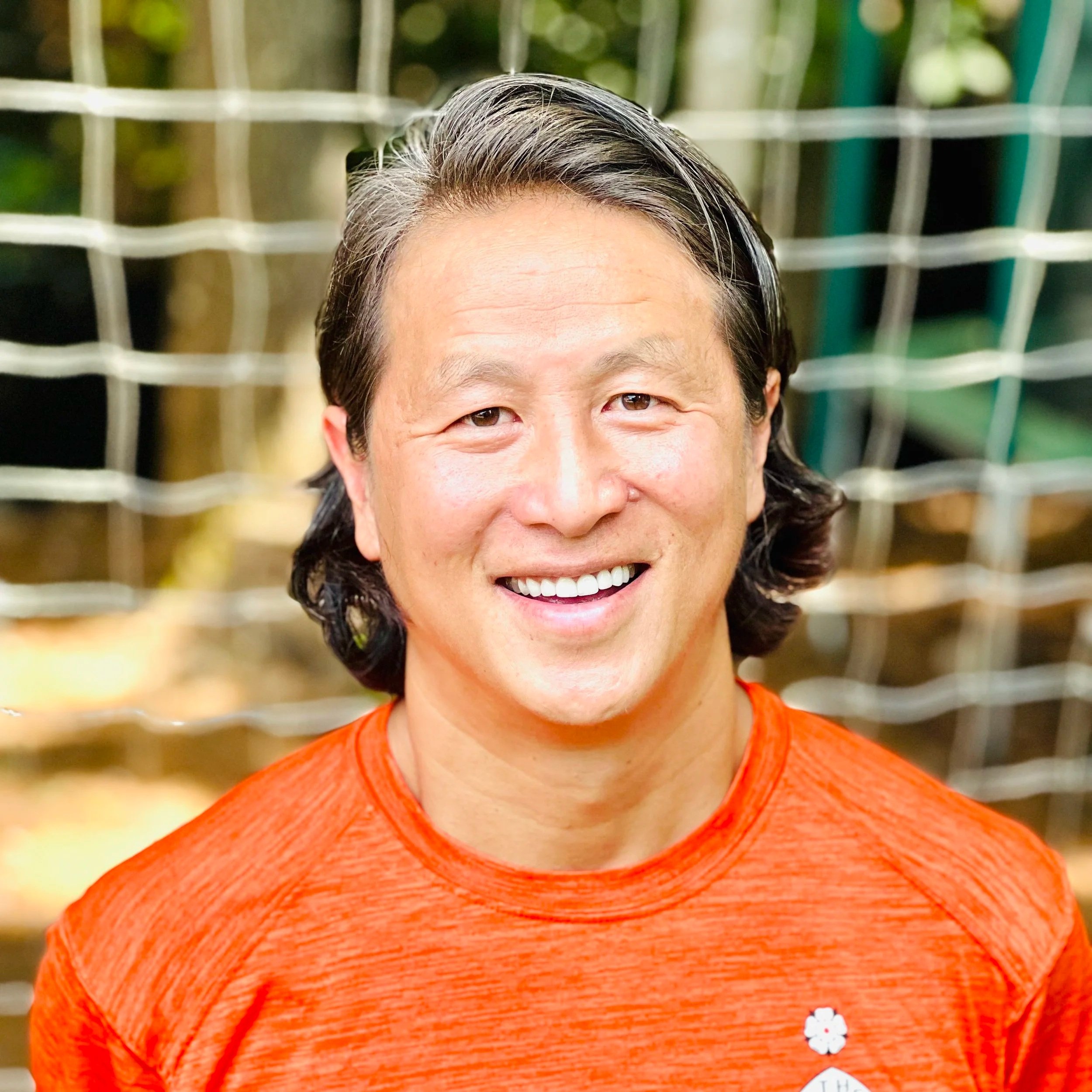 A smiling man with medium-length black hair wearing an orange athletic shirt standing outdoors with a blurry background of greenery and a white fence.