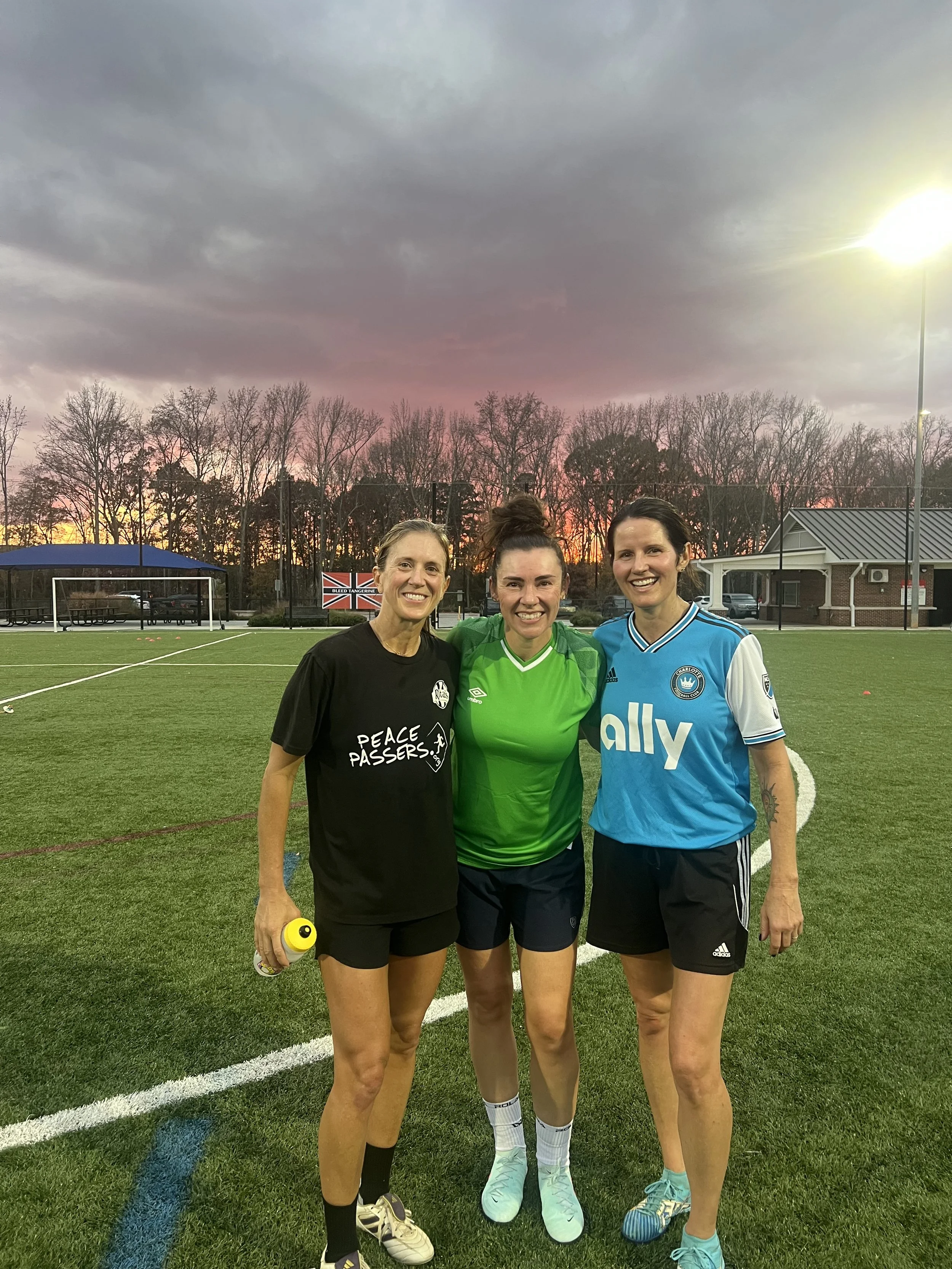 Three women standing on a soccer field at sunset, smiling, with trees and a small building in the background, dressed in sports attire.