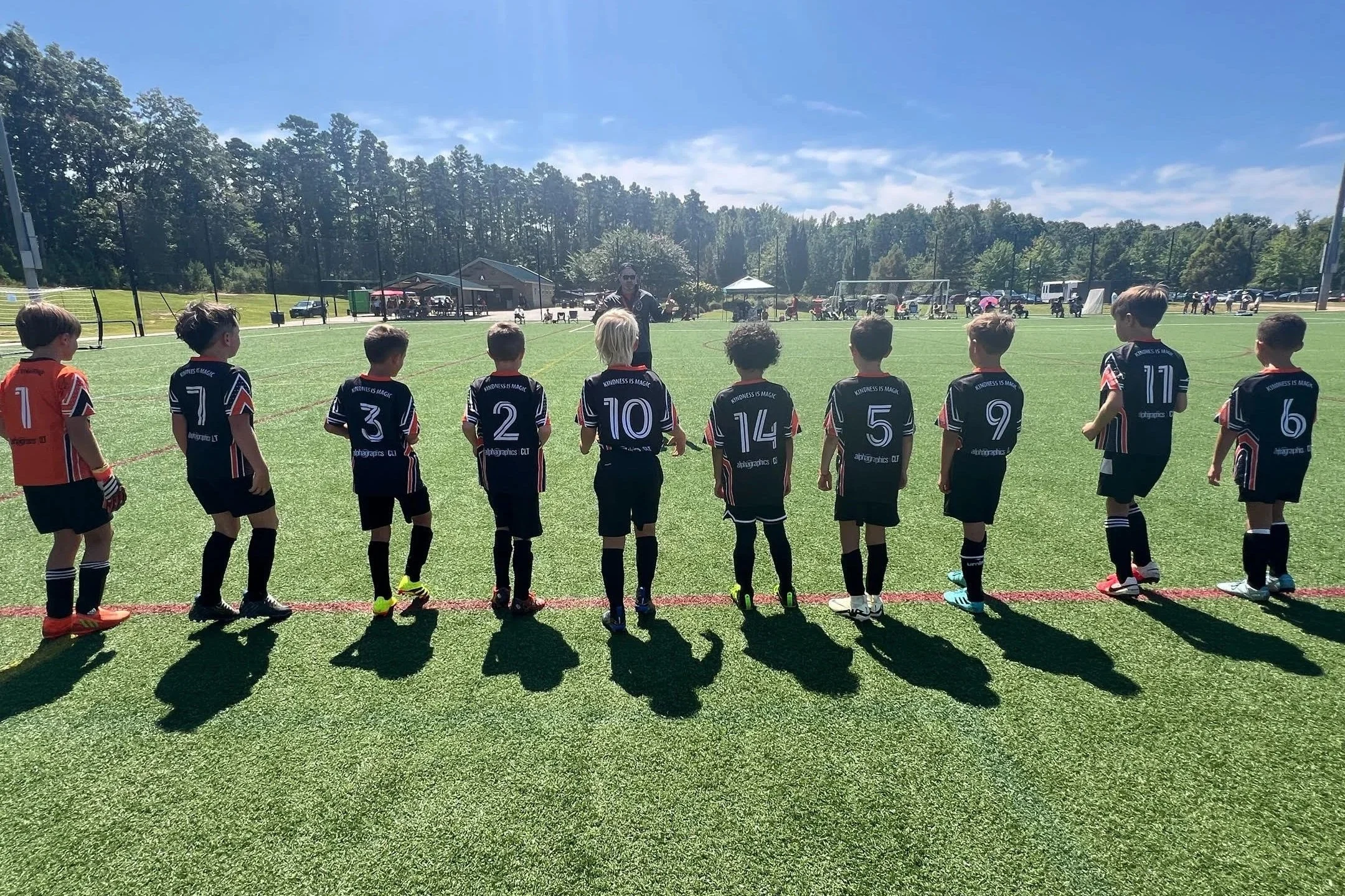 Young soccer players in black and red jerseys standing in a line on a soccer field, facing a coach.