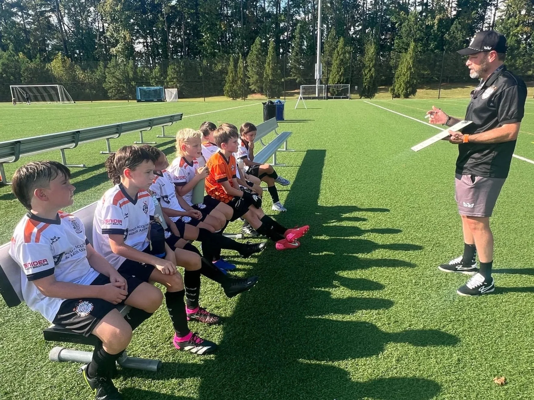 A coach giving instructions to a youth soccer team seated on a bench on a soccer field with green grass and trees in the background.