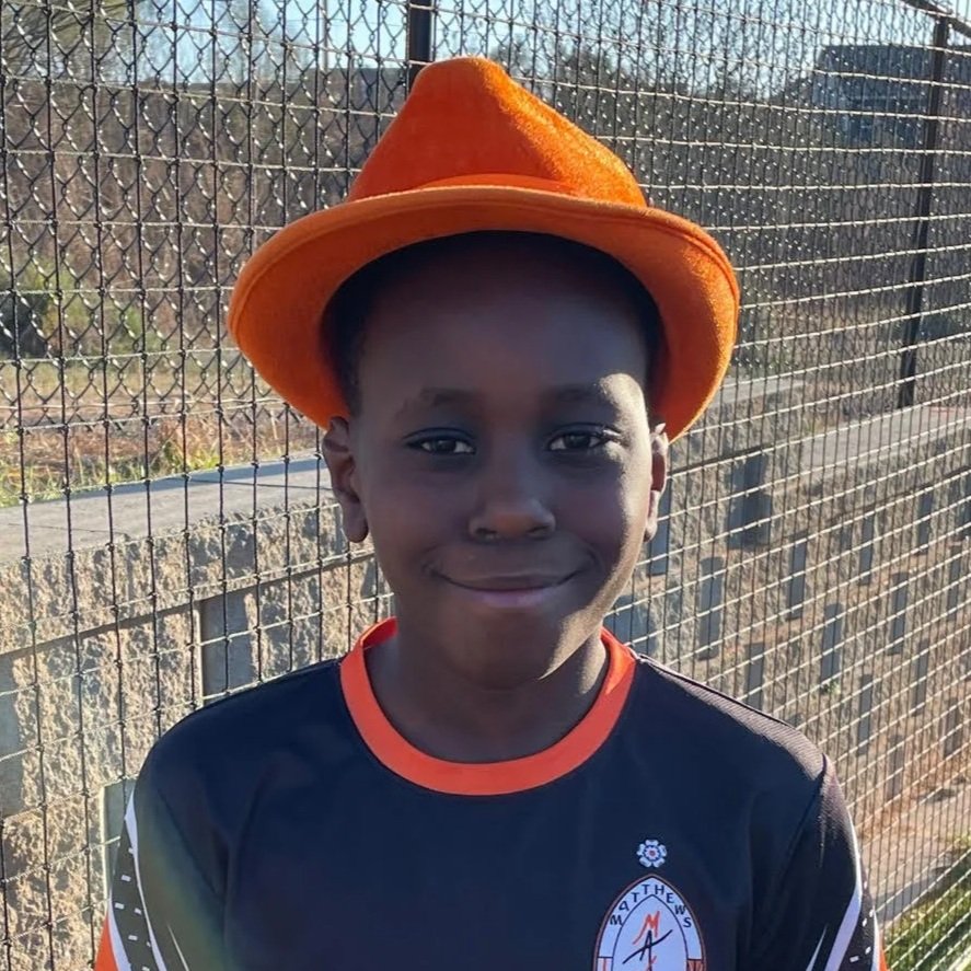 A young boy wearing an orange hat and black sports jersey, standing outdoors in front of a metal fence.