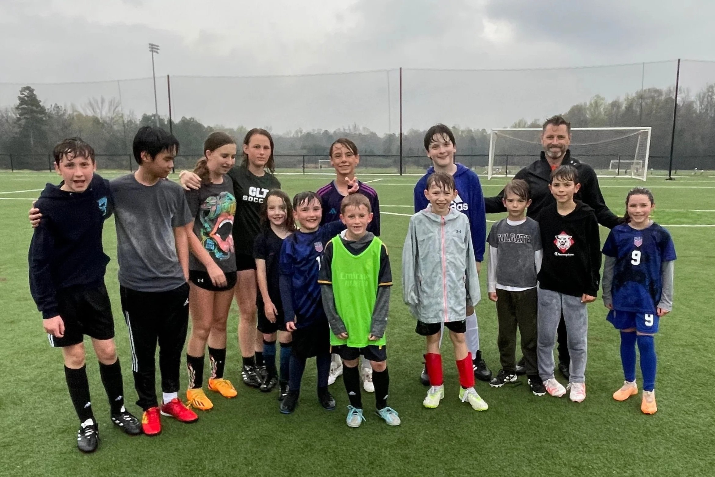 A group of children and a coach standing on a soccer field after a game or practice, some wearing soccer jerseys and athletic clothing, with a soccer goal and field in the background.