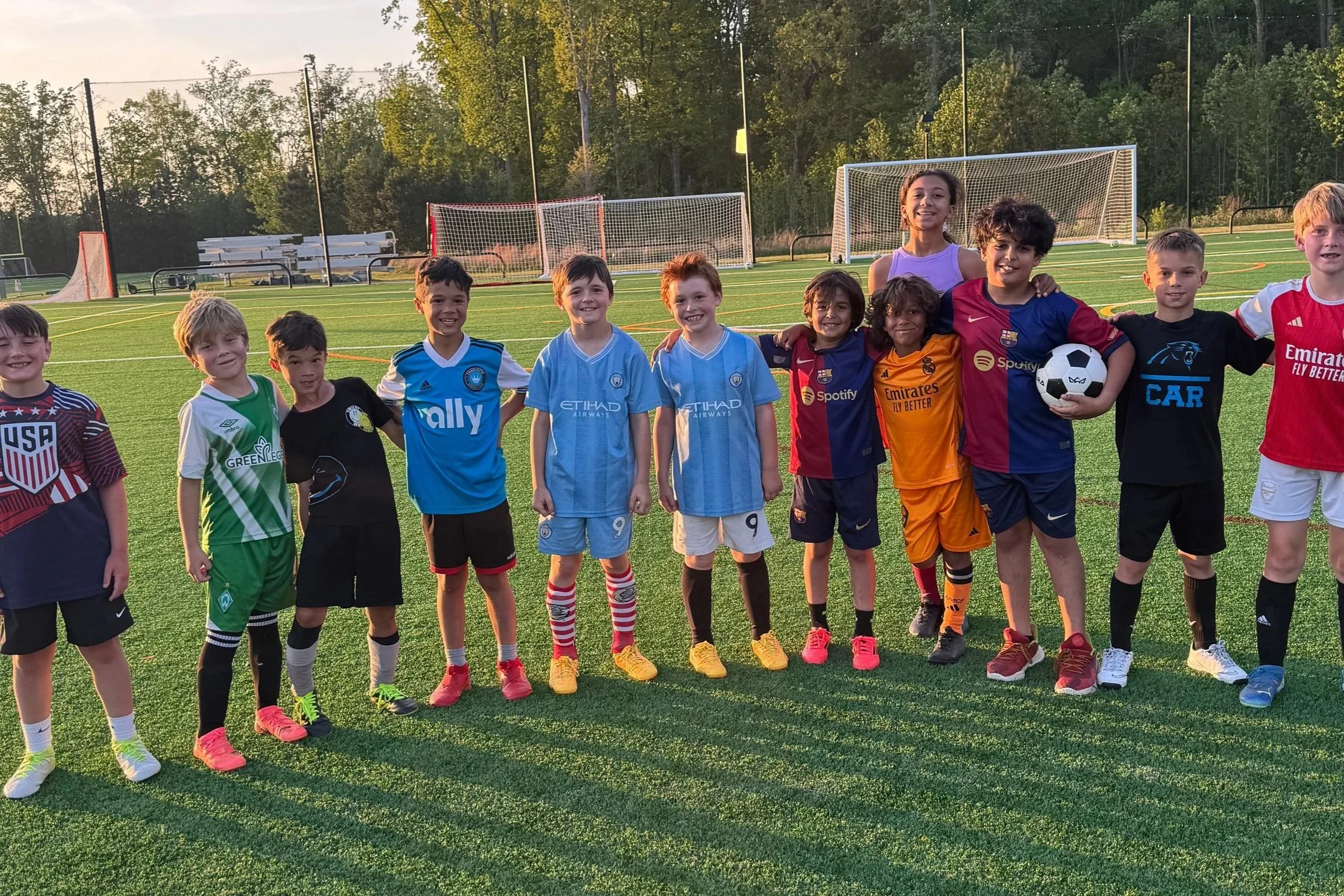 Group of diverse children and two adults, smiling on a soccer field during sunset, wearing sports jerseys, with soccer goals in the background.