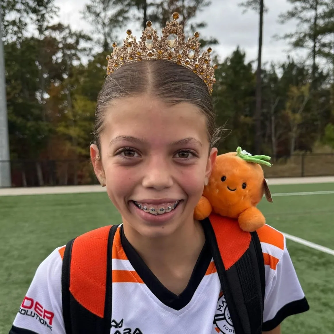A young girl wearing a tiara and a sports jersey, smiling with braces, carrying an orange plush toy on her shoulder, on a sports field with trees in the background.