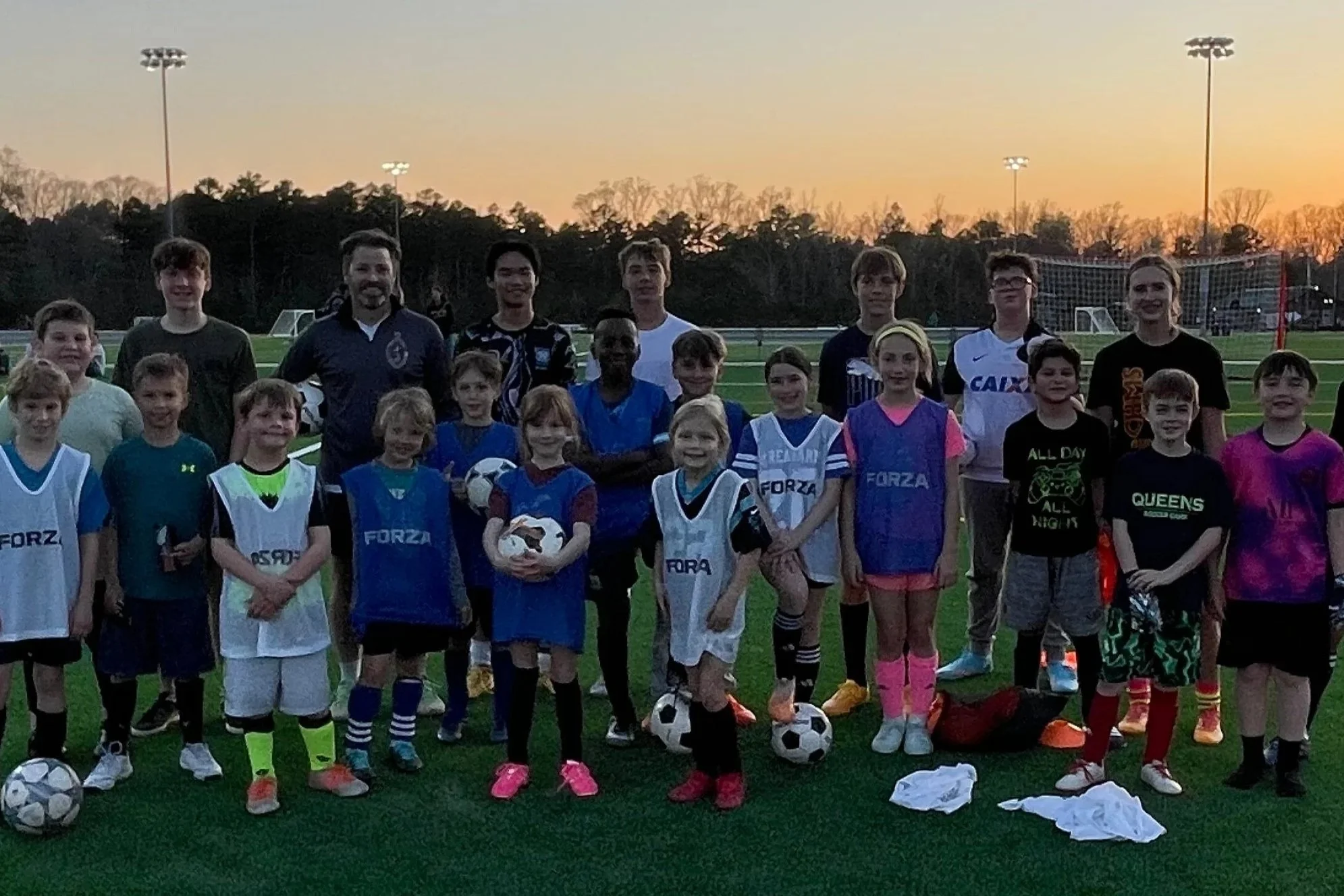A group of children and adults on a soccer field at sunset, some children wearing sports jerseys and holding soccer balls.