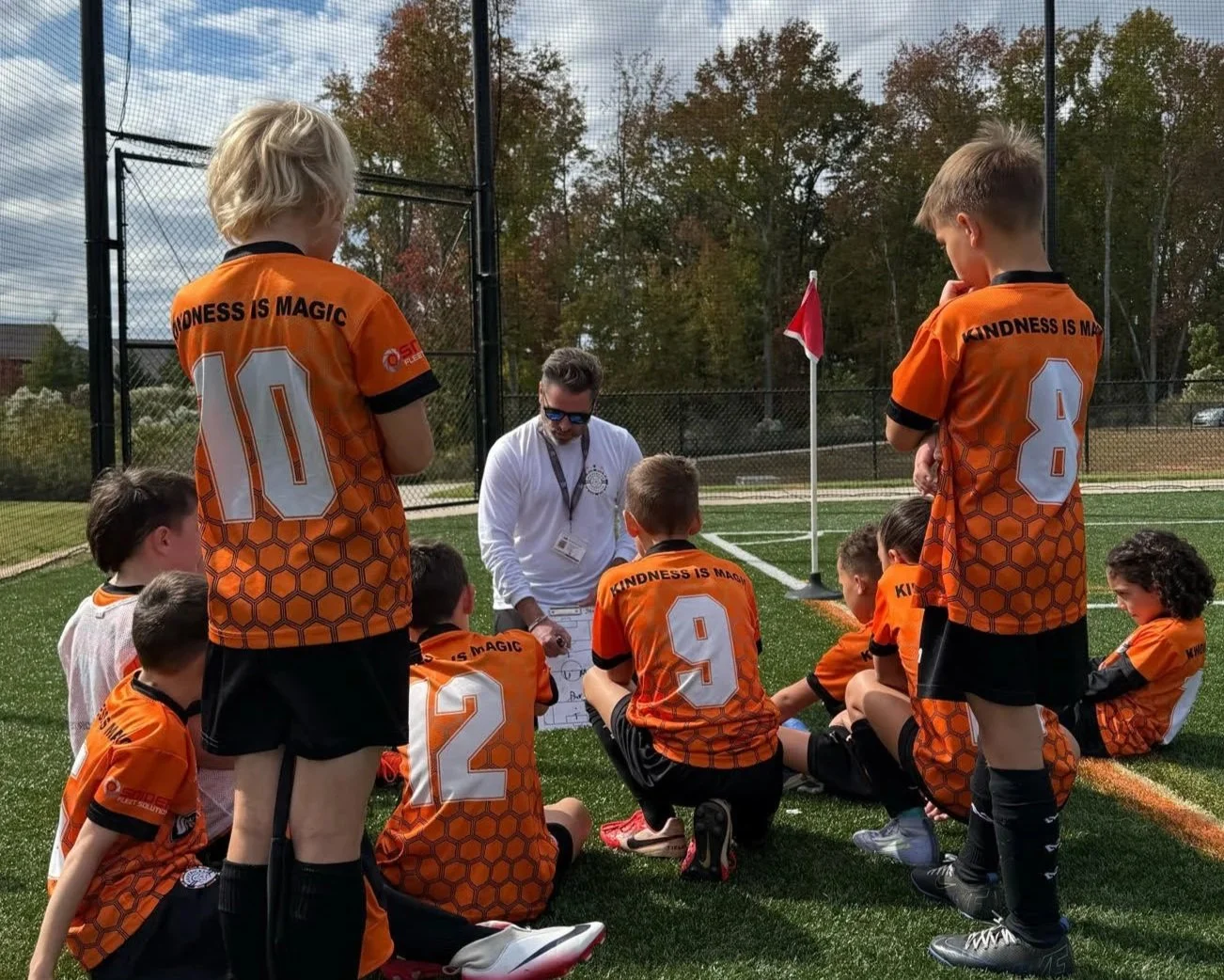 Kids in orange soccer uniforms sitting on the field in a huddle listening to coach during practice or game on a sunny day.