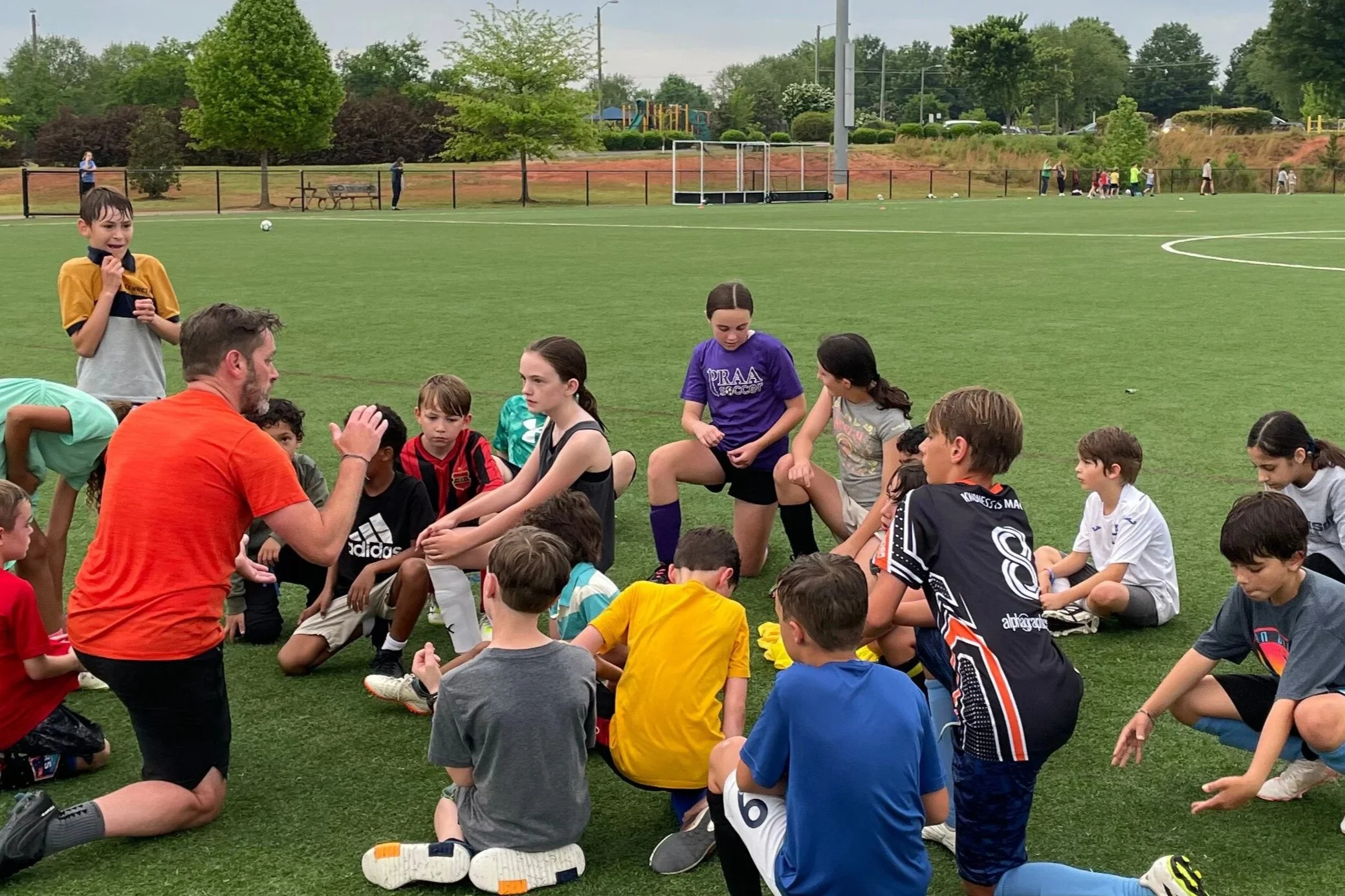 A coach giving instructions to a group of children sitting on a soccer field during practice, with some children listening attentively and others have their hand raised.