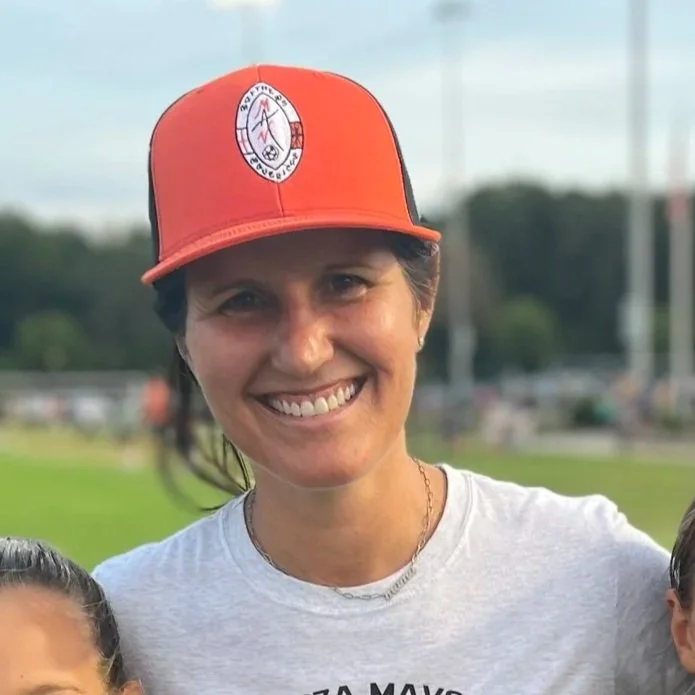 Woman smiling wearing an orange baseball cap and a white shirt outdoors.