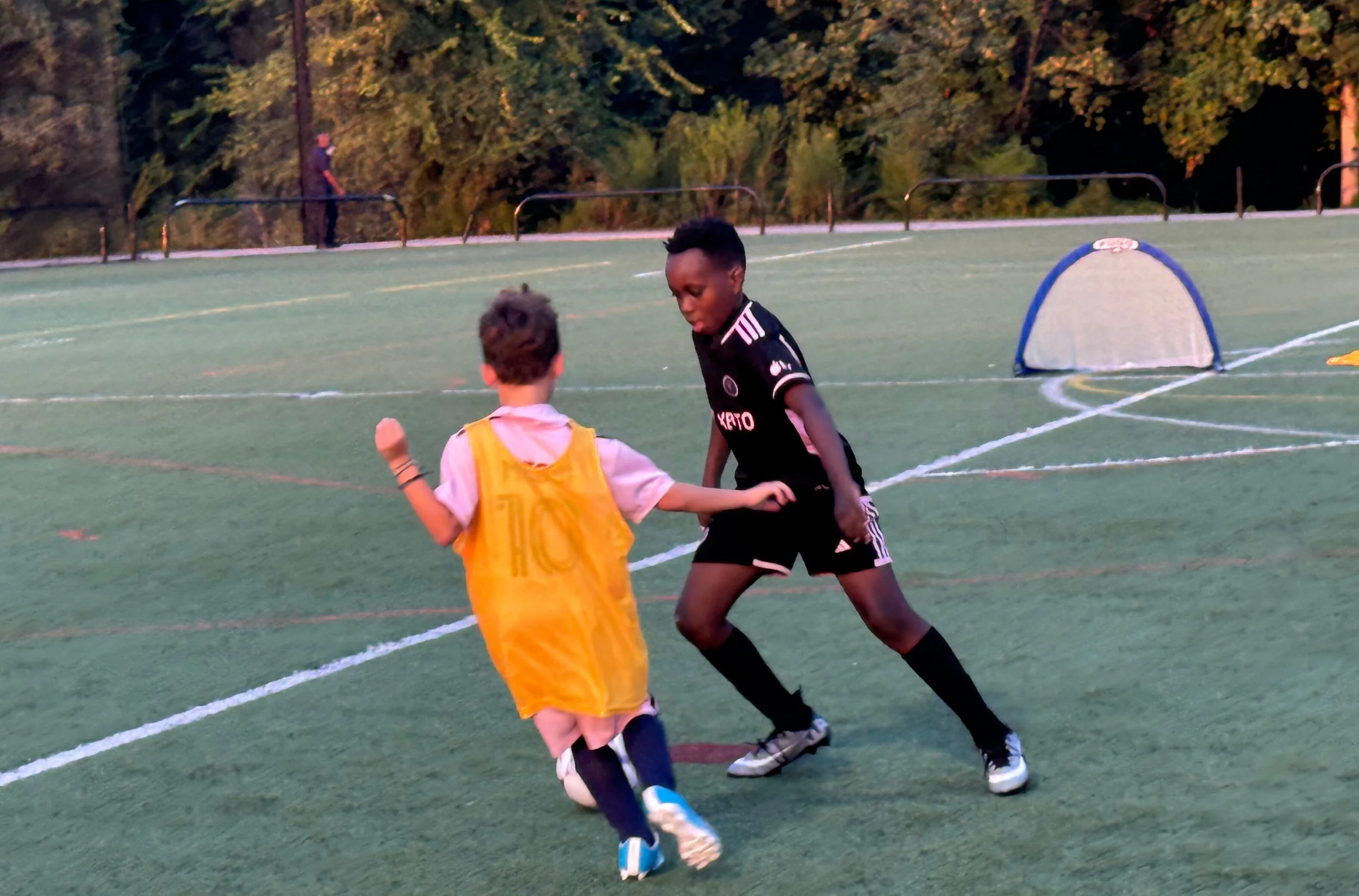 Two young boys playing soccer on an outdoor field, one wearing a yellow jersey and the other in a black sports outfit, with small soccer goals and trees in the background.