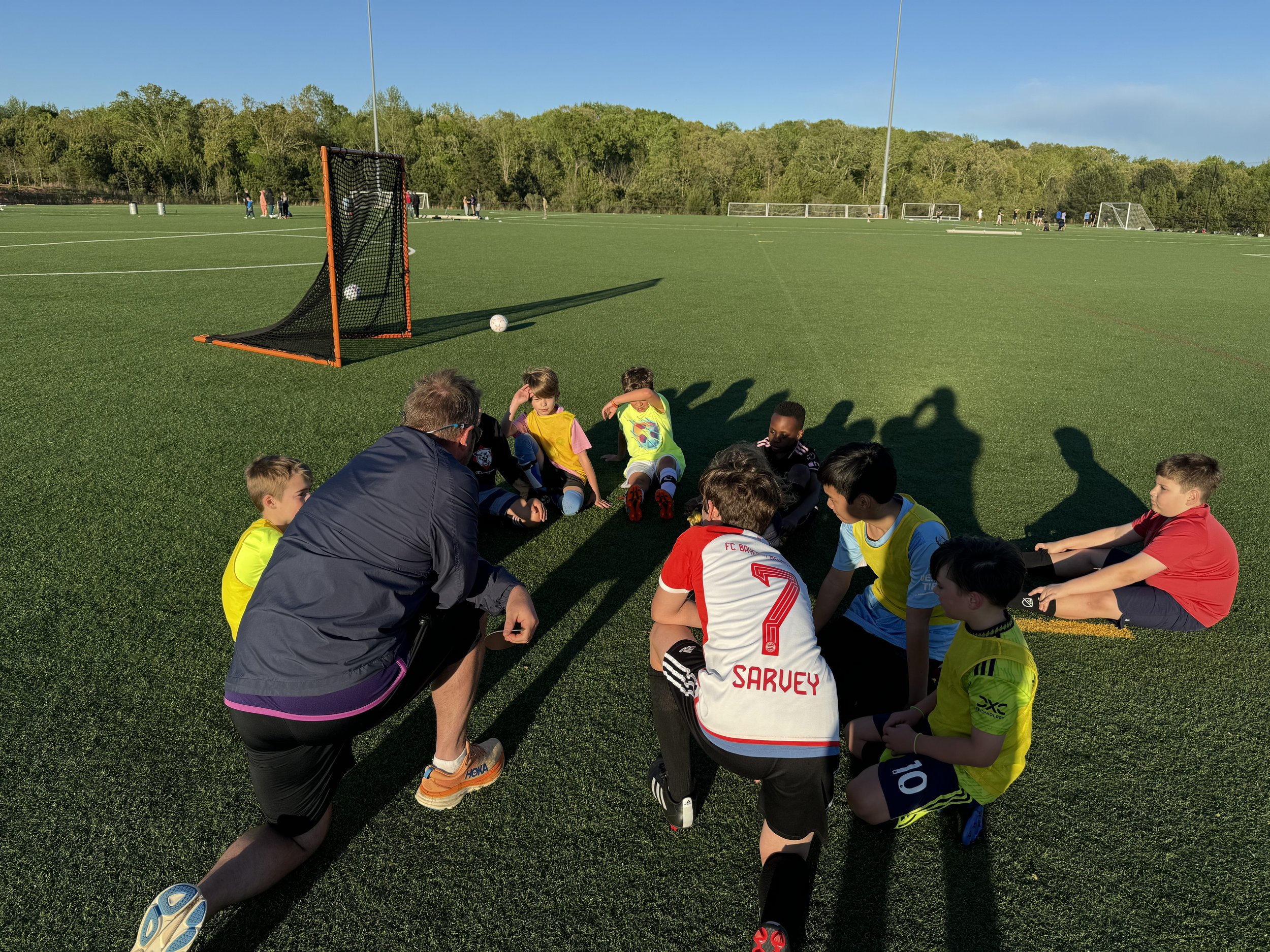 A group of children sitting on a soccer field with a coach, during a soccer practice or game, with goal nets and trees in the background.