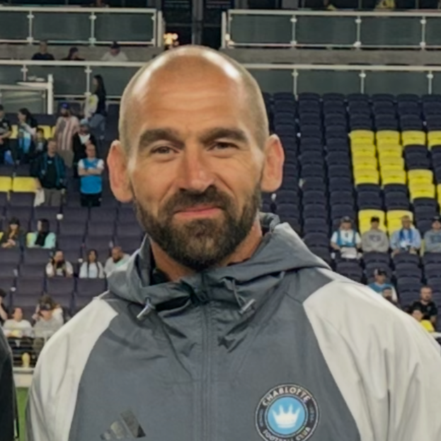 A man with a beard smiling, wearing an orange sports jersey, standing outdoors with trees in the background.