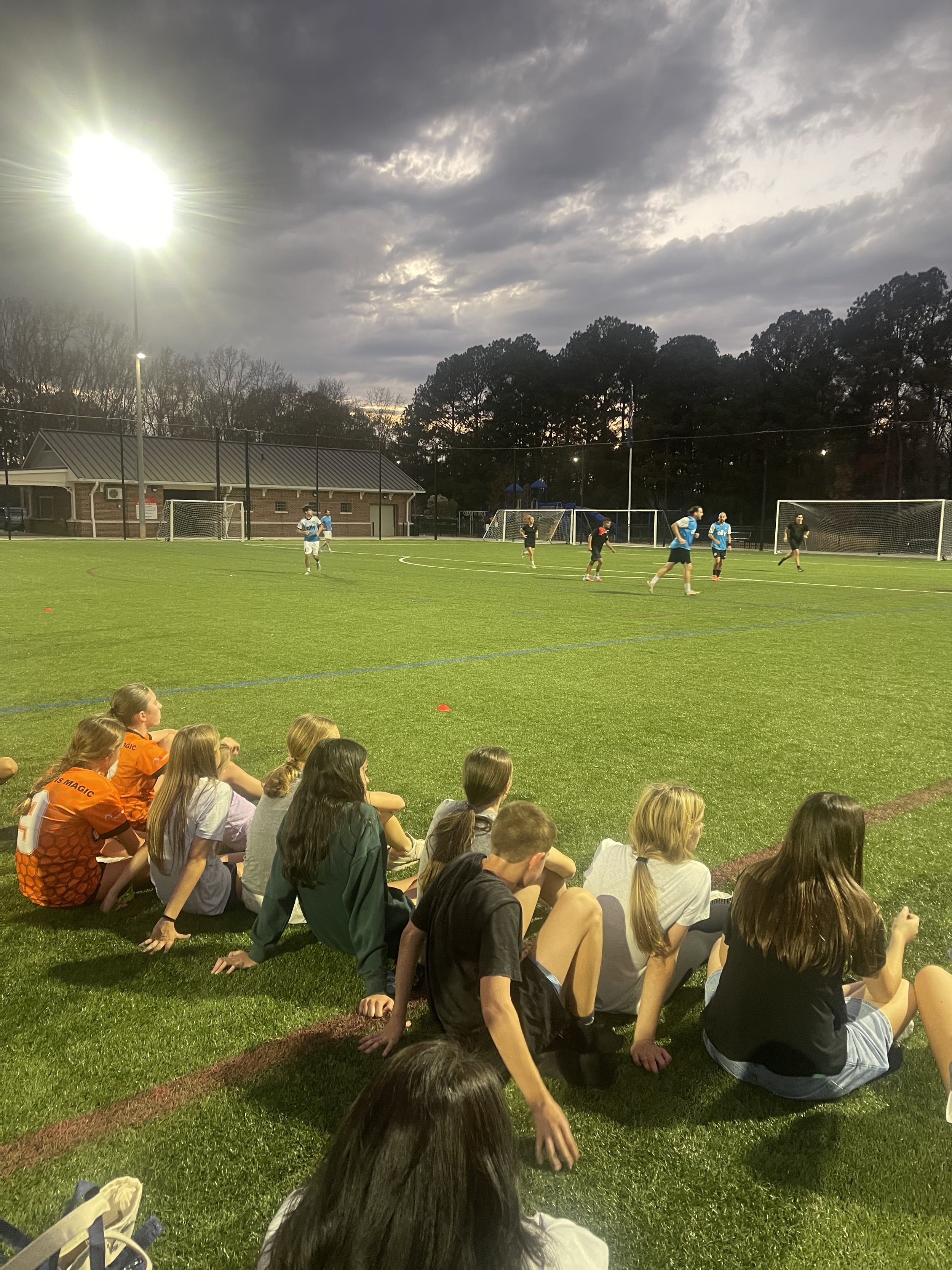 Group of children seated on the grass watching a soccer practice or game on a field under a cloudy evening sky.