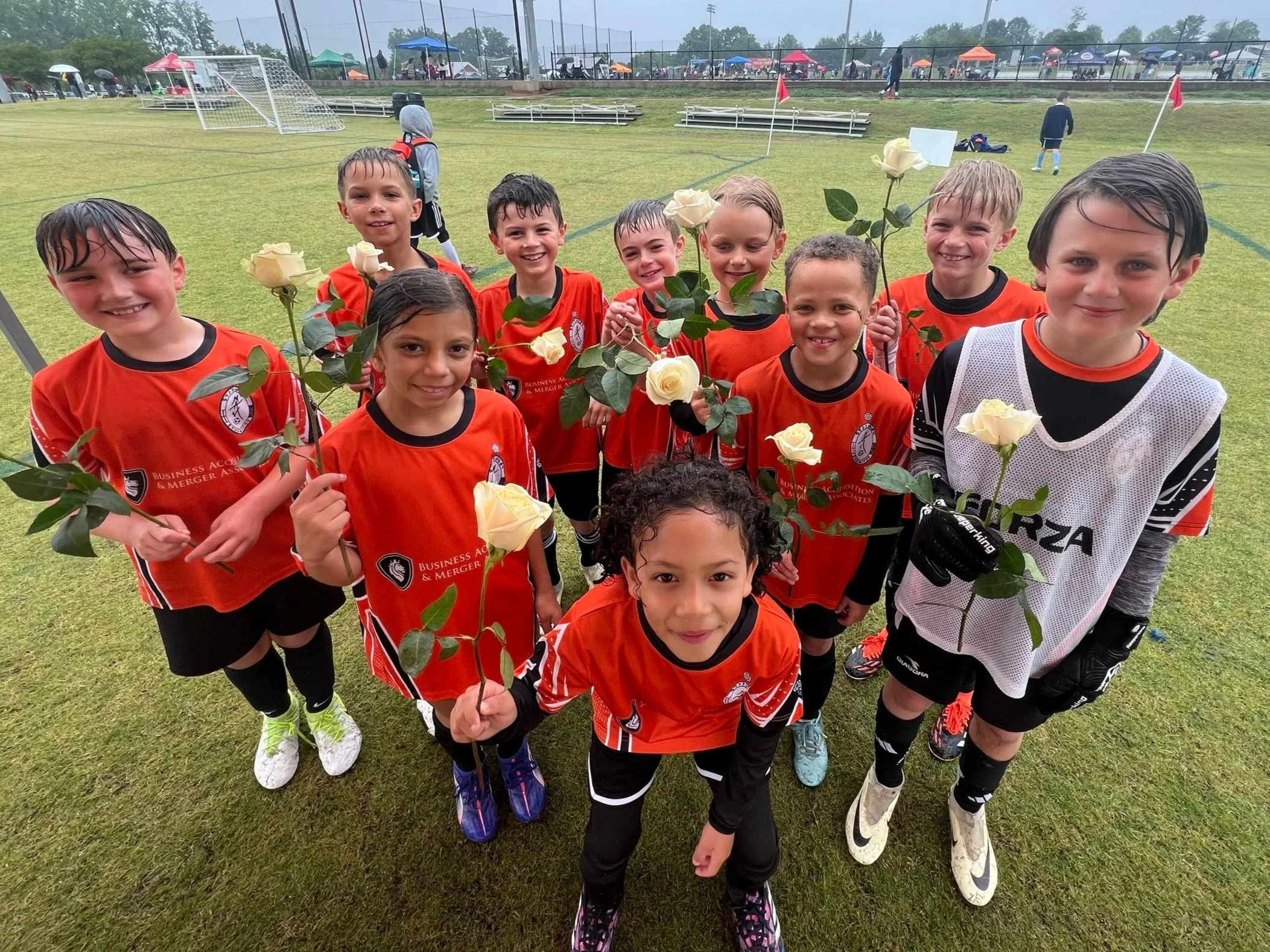 A group of young children in soccer uniforms holding white roses on a soccer field after a game in the rain.