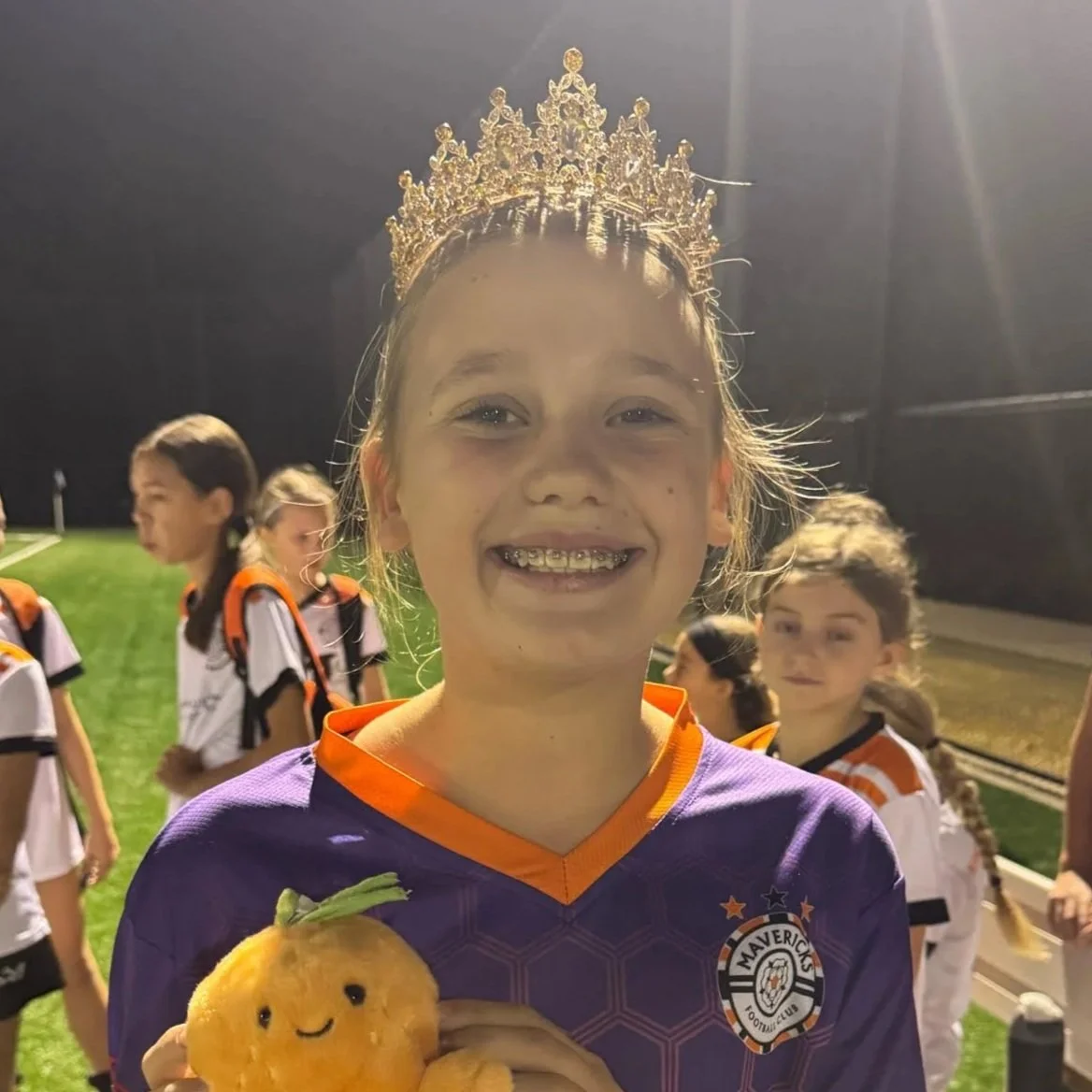 A young girl wearing a purple soccer jersey and a gold crown, holding a plush toy, smiling at night on a soccer field with other children in sports uniforms in the background.