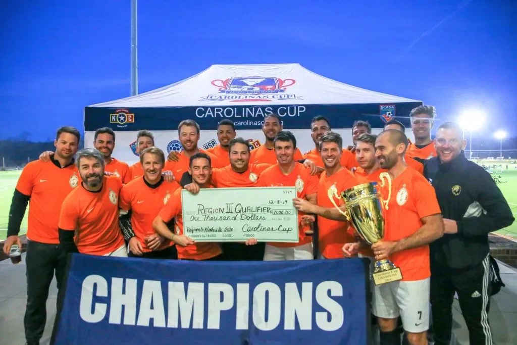Soccer team celebrating victory at the Carolinas Cup, holding a large check for $1,000 and a trophy, with a banner that reads 'Champions' and a tent behind them.