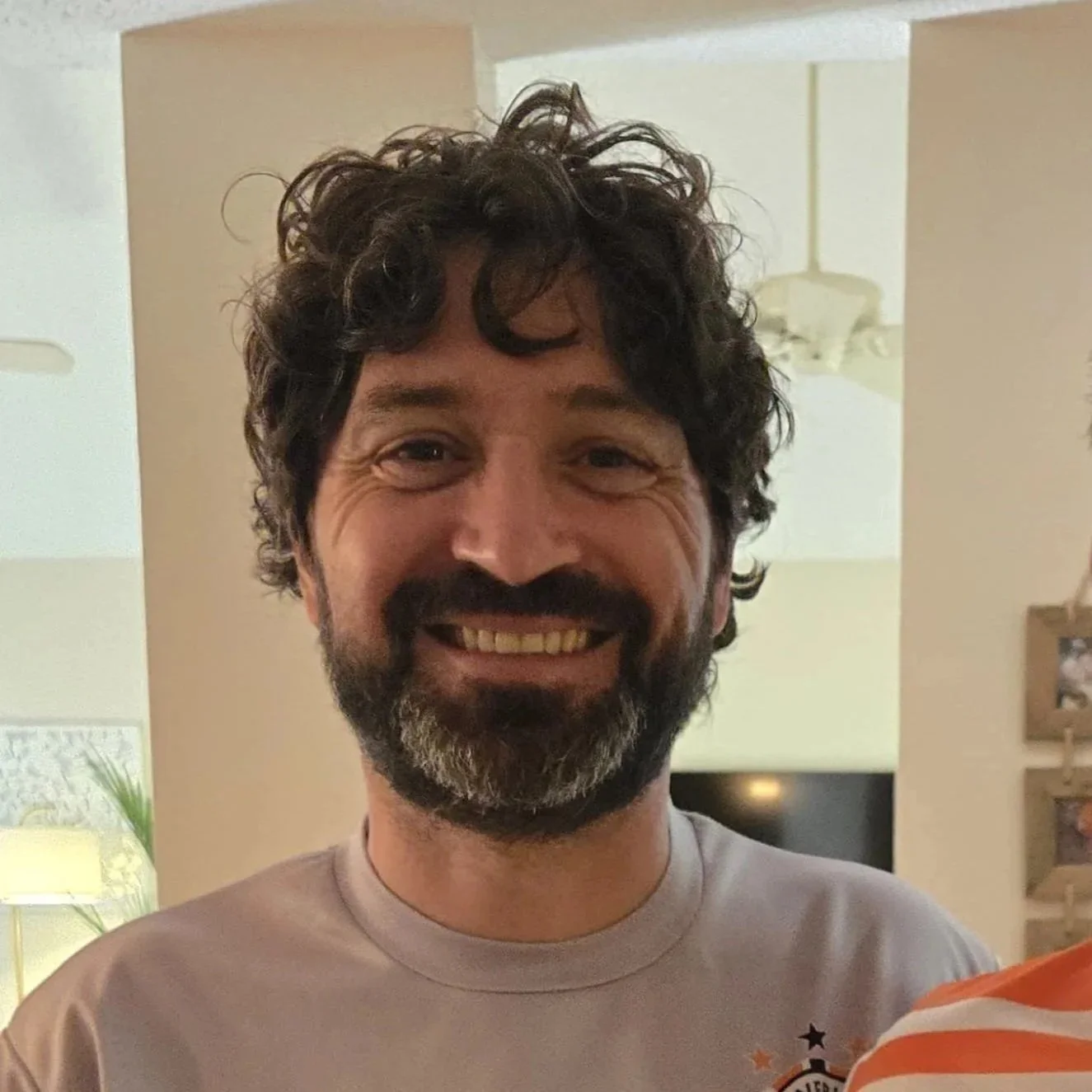 A smiling man with curly dark hair and a beard, standing indoors with a beige wall and ceiling fan in the background, wearing a light gray shirt.
