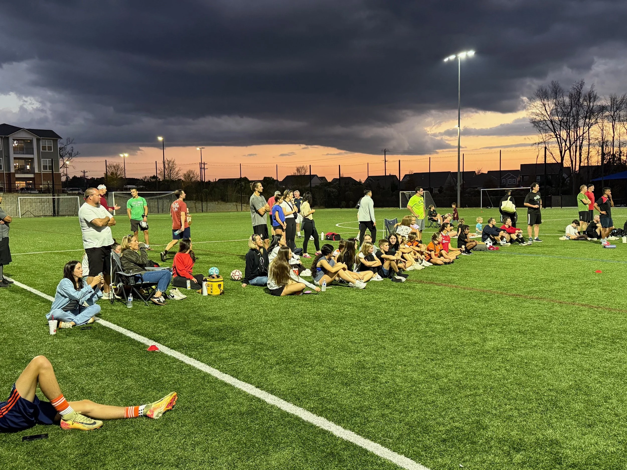 A group of people, including children and adults, sitting and standing on the sideline of a soccer field during sunset under dark clouds, watching a soccer game.
