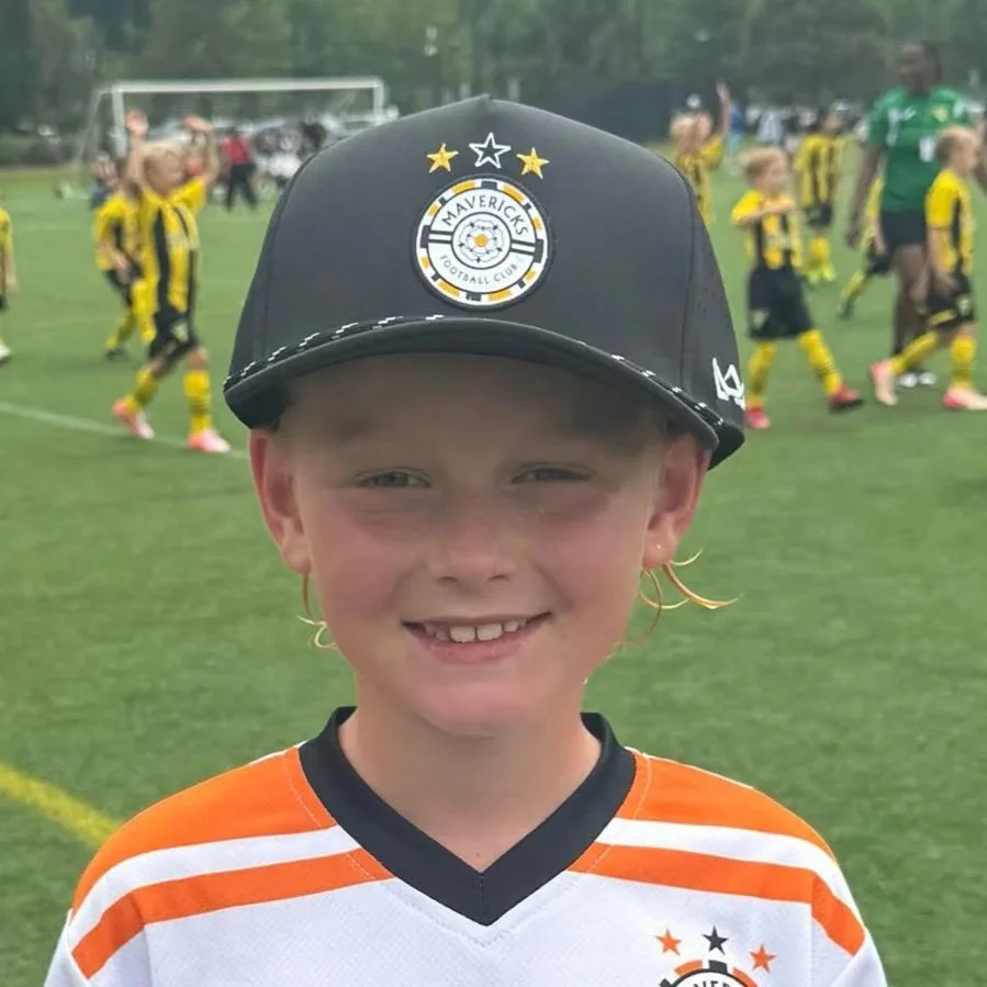 Young boy smiling at a soccer game, wearing an orange and white jersey and a black Maverick's Football Club cap, with a soccer field and players in yellow and black uniforms in the background.