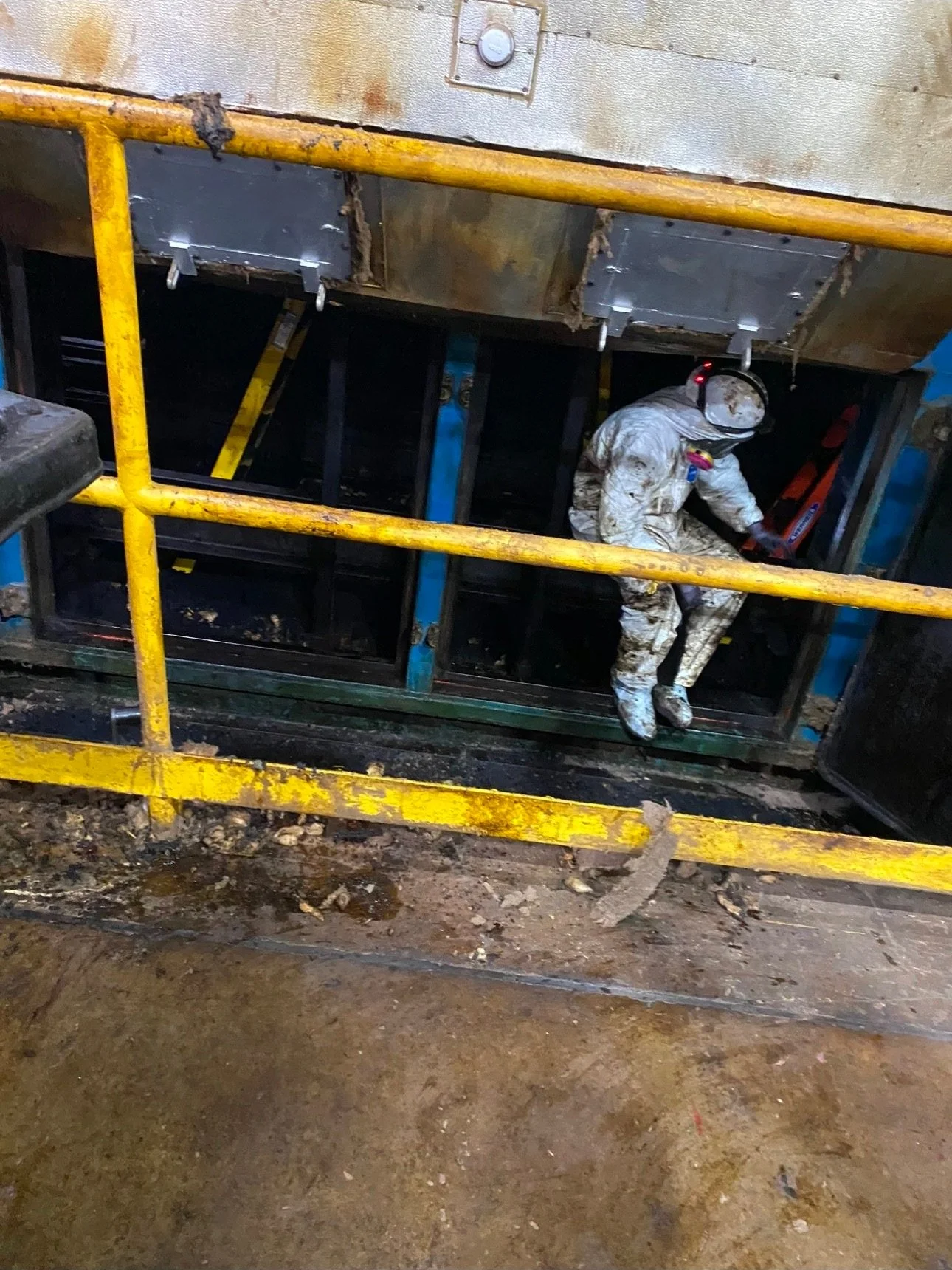 Worker wearing a white protective suit, helmet, and safety goggles working underneath a large piece of industrial equipment.