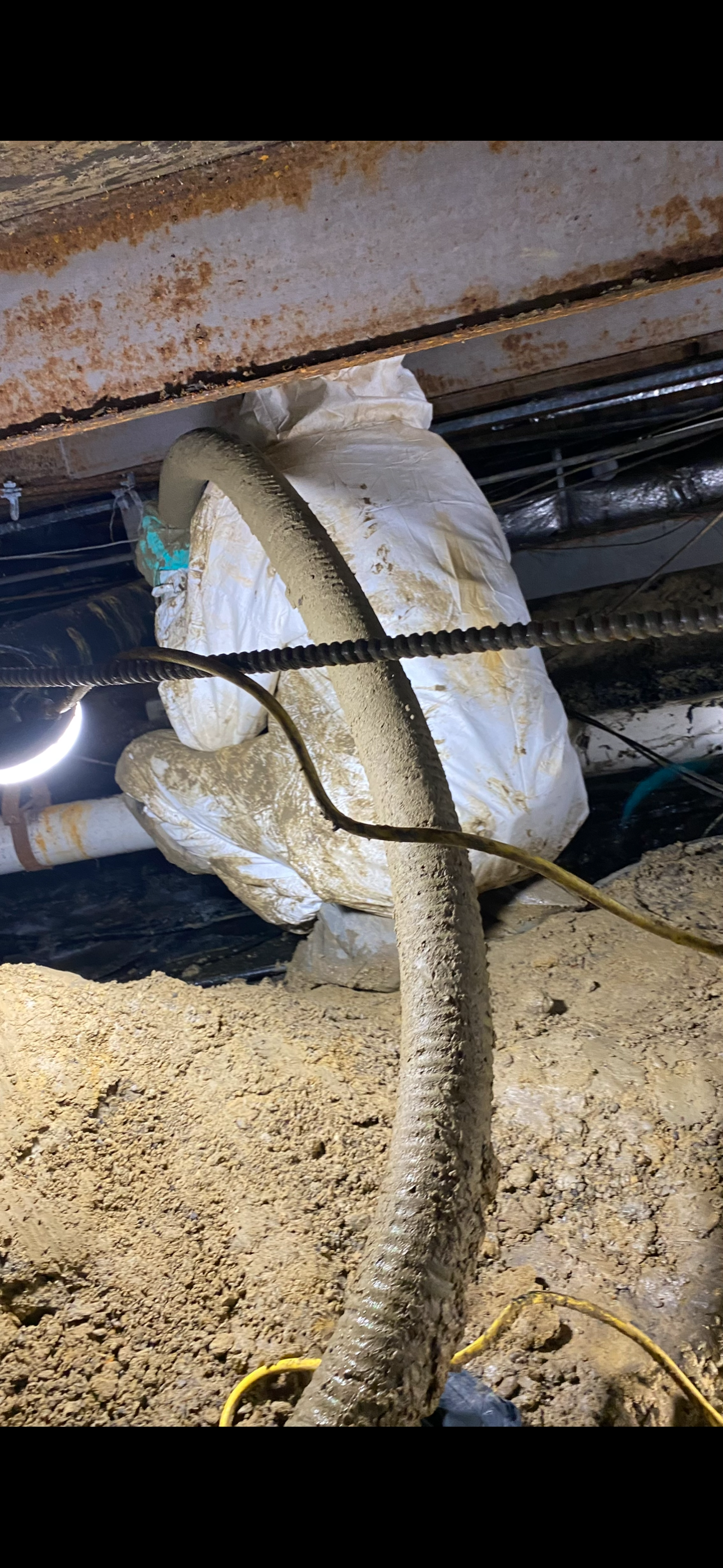 A construction site image showing a dirty hose, a worker in a white protective suit, and exposed pipes and rebar in an excavated area.