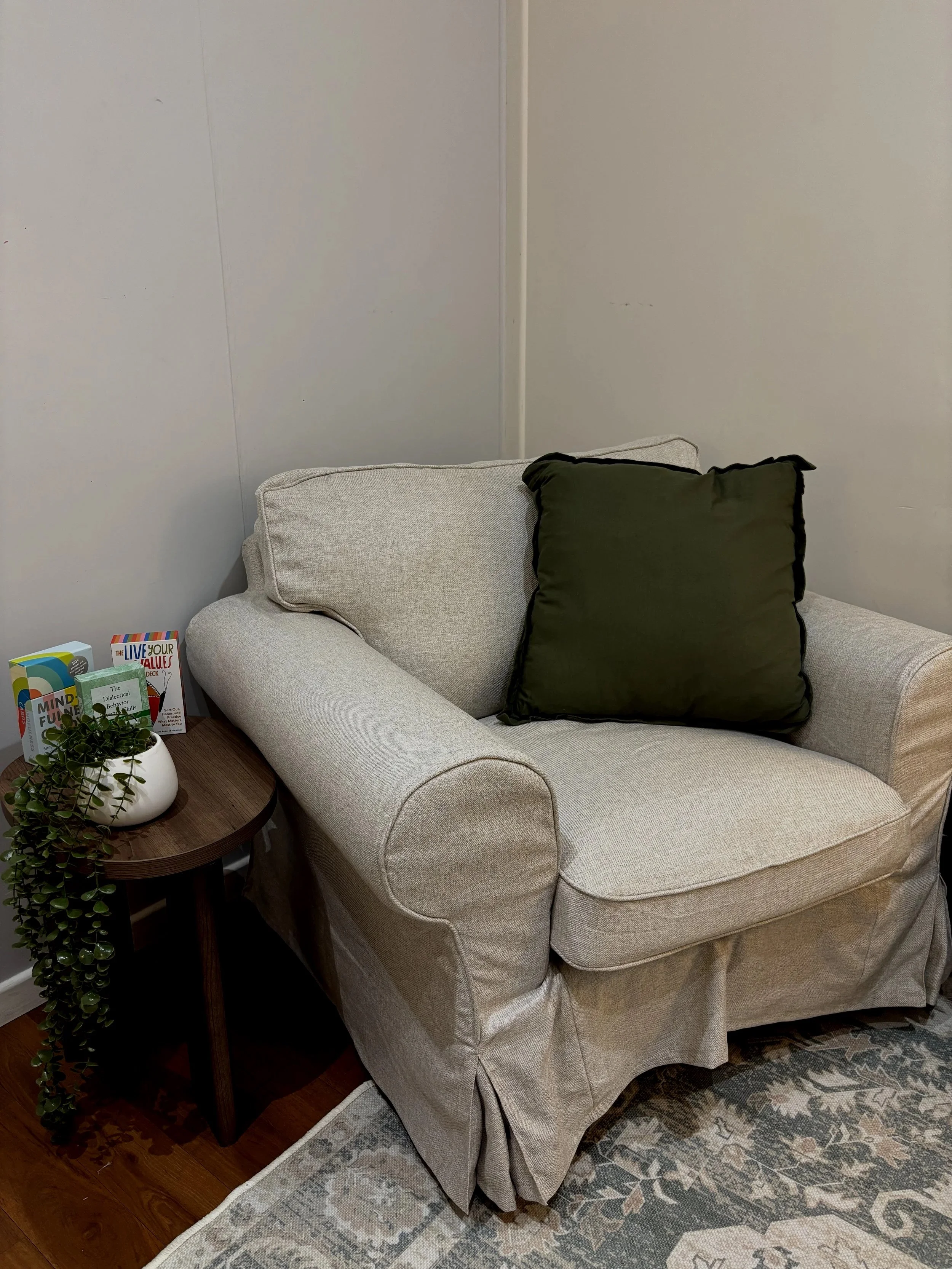 A beige armchair with a dark green pillow, a small wooden side table, and a potted plant with books next to it, in a cozy corner with a patterned rug.