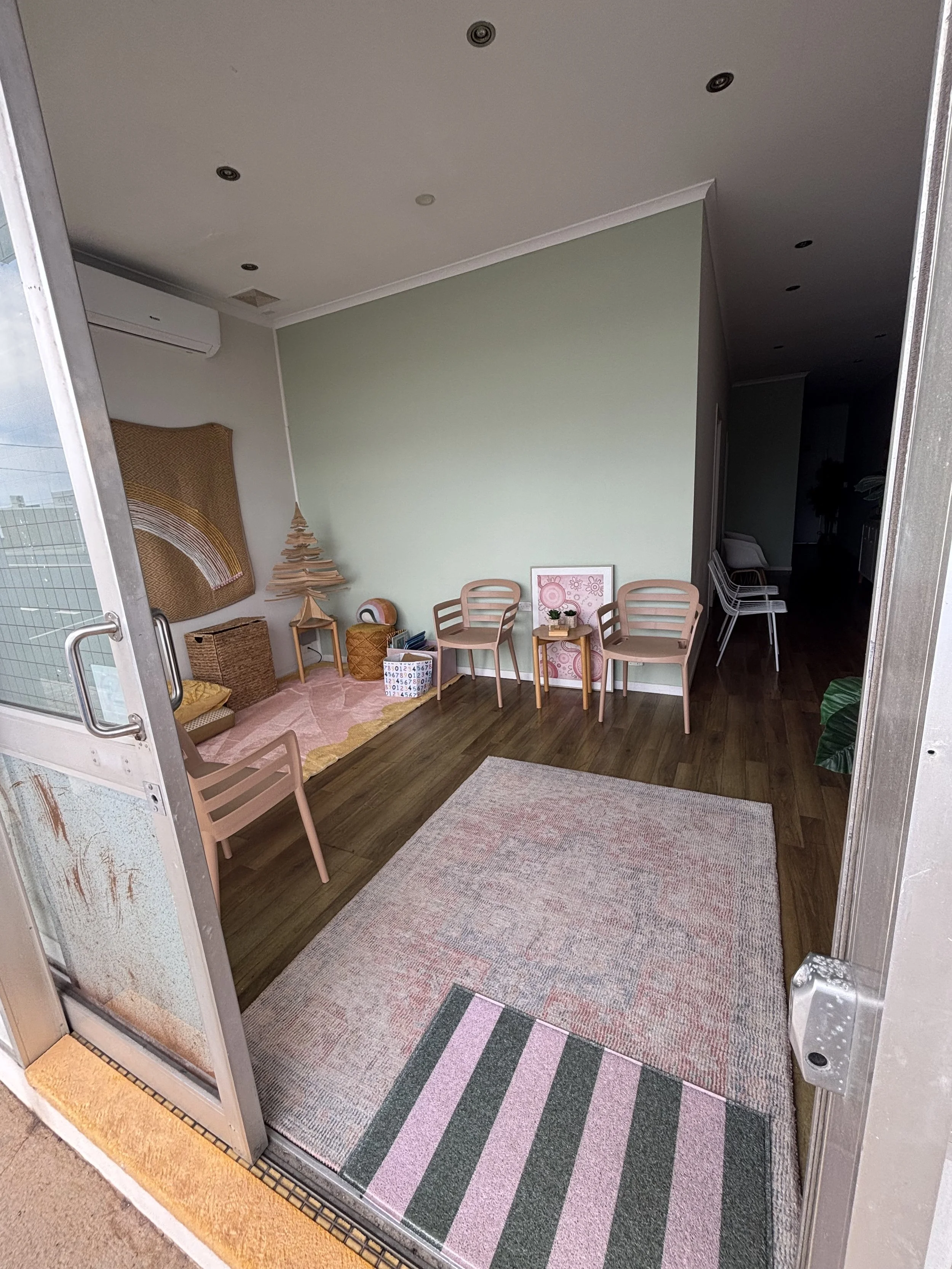 View of a cozy, decorated indoor space with a light green wall, wooden chairs, small tables, a pink and gray striped rug, and decorative art on the walls.