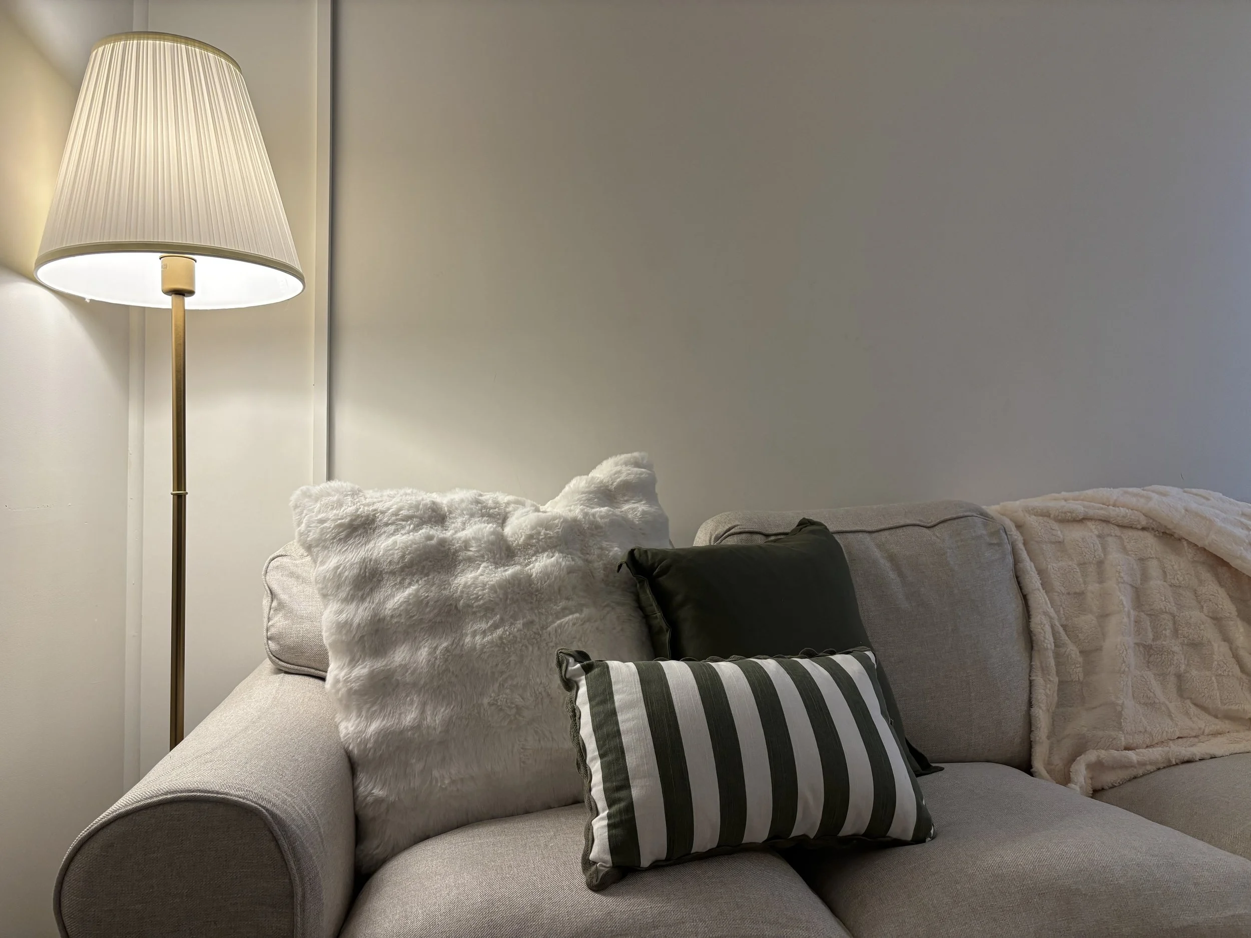 A beige sofa with multiple pillows, including a white furry pillow, a black pillow, and a black-and-white striped pillow. There is a beige blanket draped over the sofa. To the left, a tall floor lamp with a white lampshade provides lighting. The background includes a plain off-white wall.