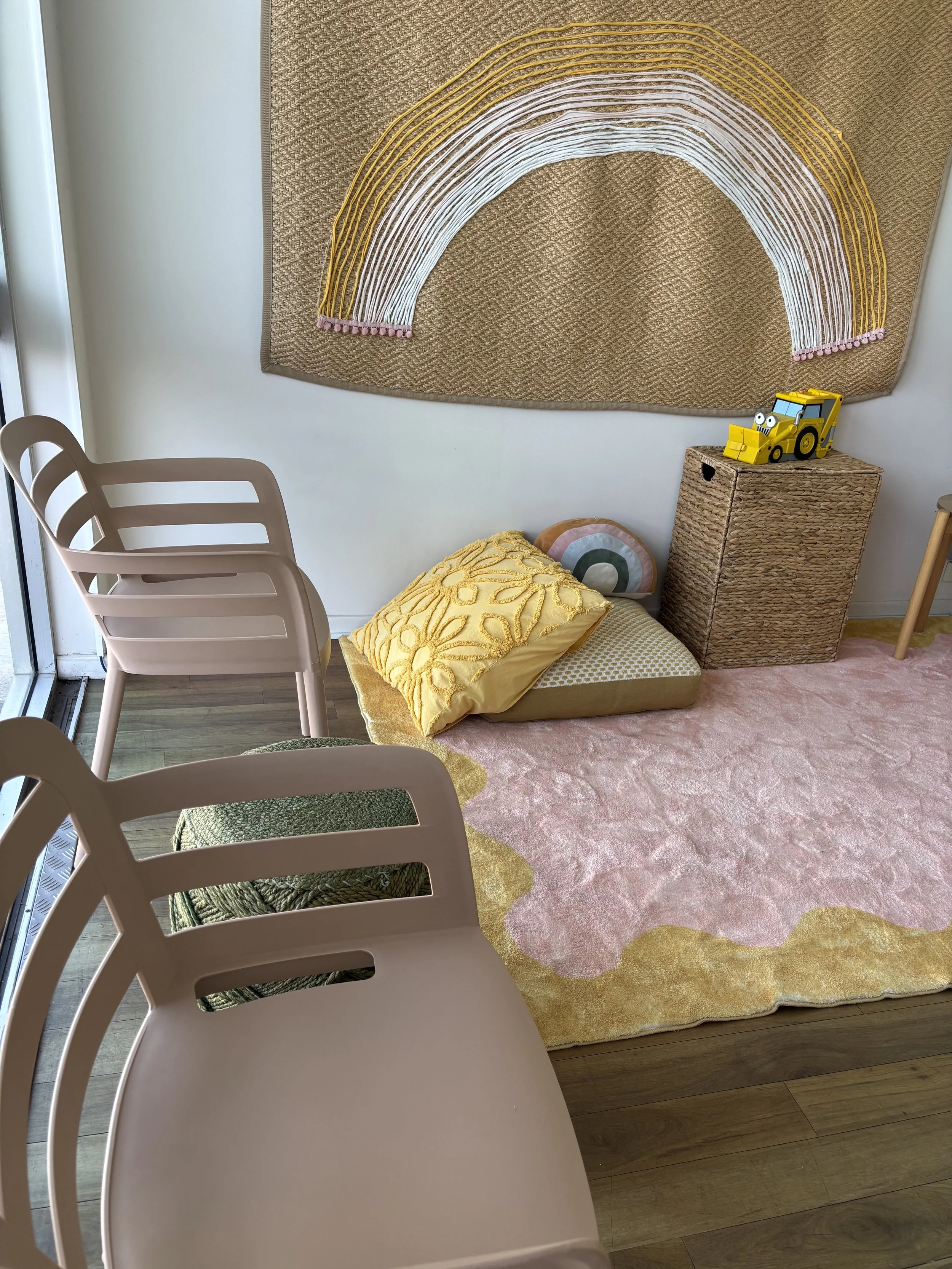 A cozy corner with a large pink and yellow rug, a pile of yellow and pastel pillows on the rug, a woven basket, a yellow toy bulldozer with googly eyes, a woven wall hanging with a rainbow design, and pink chairs around the rug near a sliding glass door.