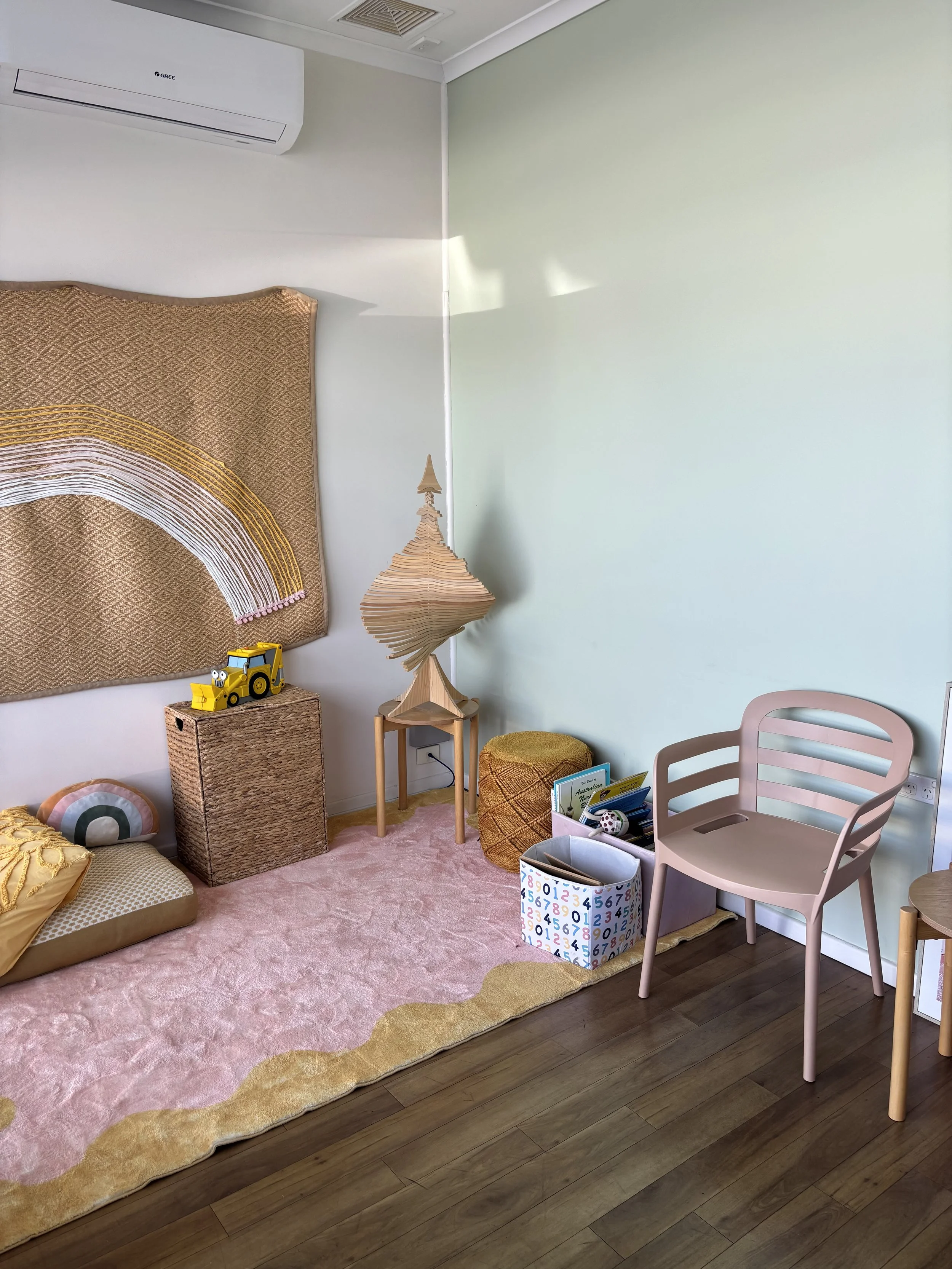 A cozy children's playroom with a pink carpet and a light green wall, decorated with plush pillows, a woven storage box, a yellow toy tractor, a textured yellow ottoman, a pastel pink chair, a small wooden table, and shelves filled with books and toys.