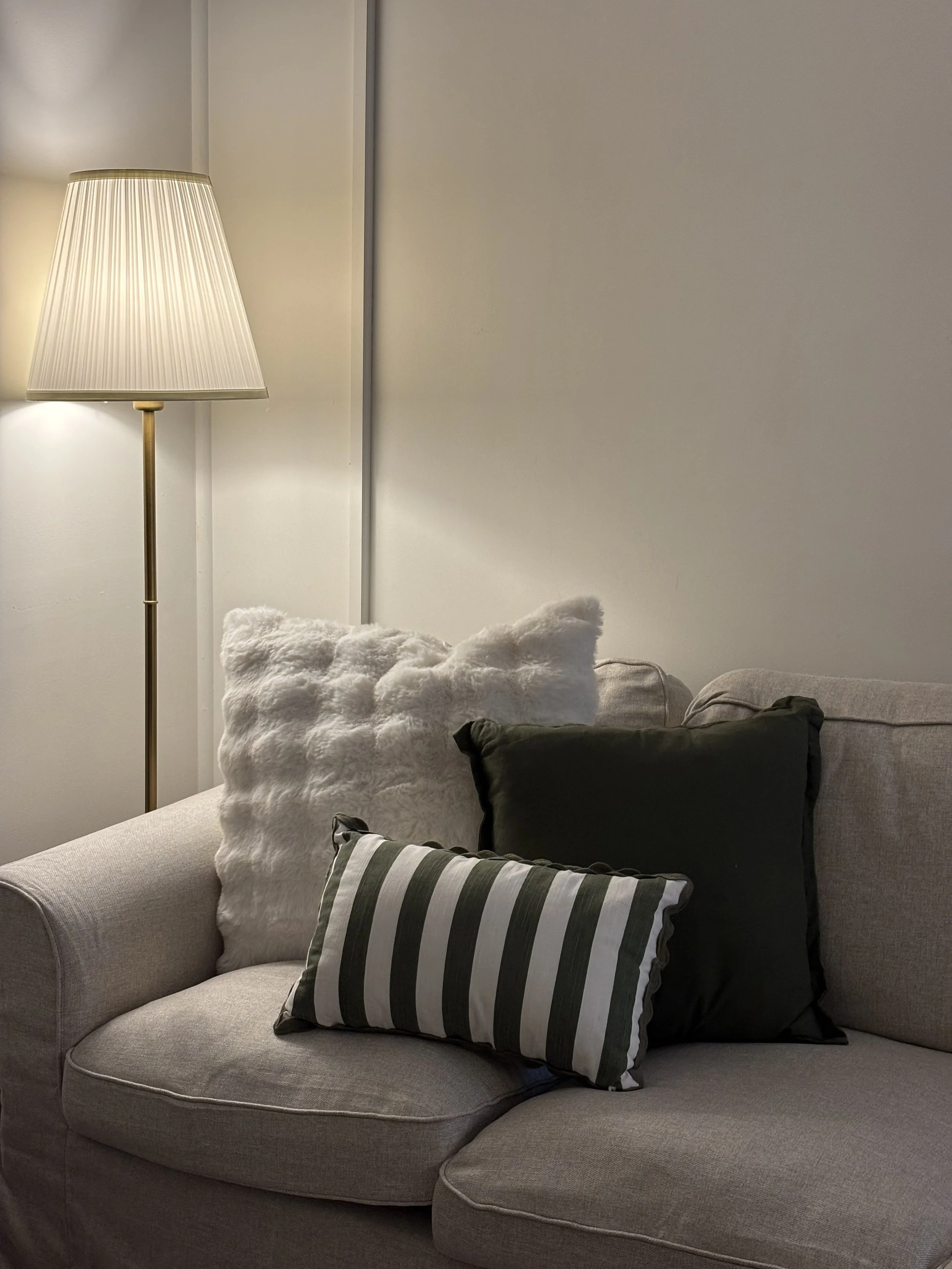 Close-up of a beige sofa with three pillows in front of a plain white wall. The pillows are plush white, dark green, and black with white and dark green stripes. To the left, there's a tall standing lamp with a beige pleated lampshade.
