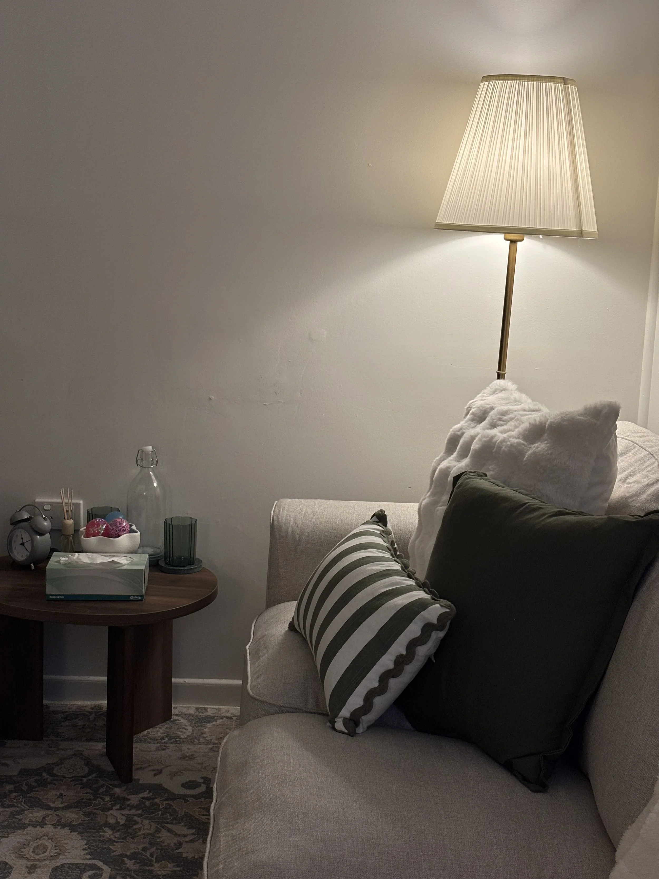 A cozy living room corner with a beige sofa, decorative pillows, a floor lamp, and a wooden side table with a tissue box, a clock, a glass bottle, and candles.