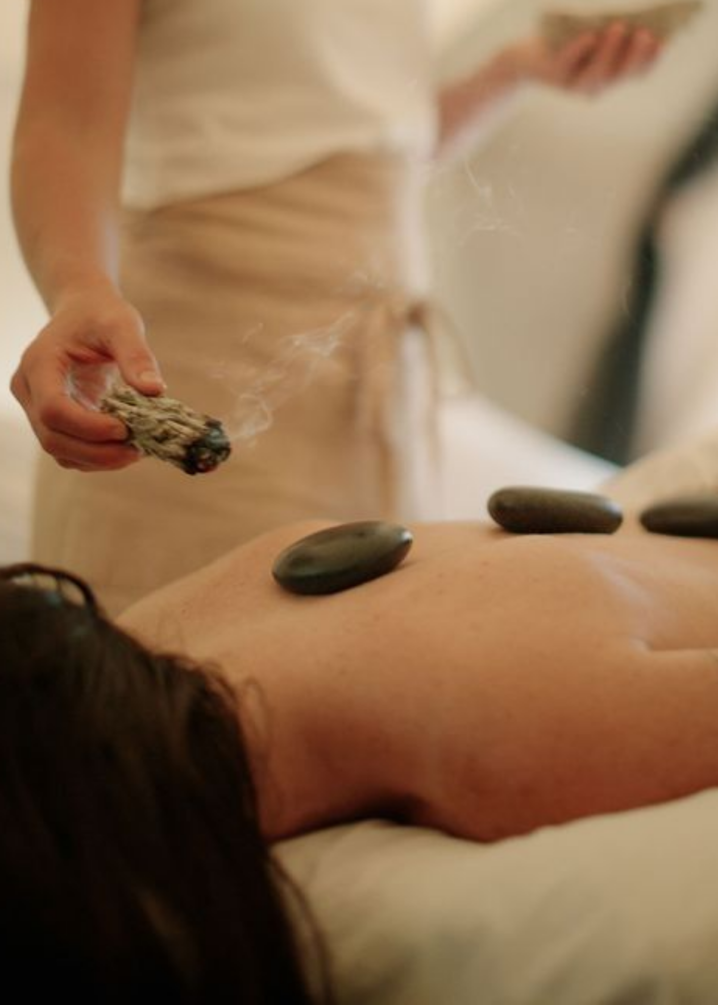 Person receiving hot stone massage with heated stones on back, and therapist holding smoldering sage.