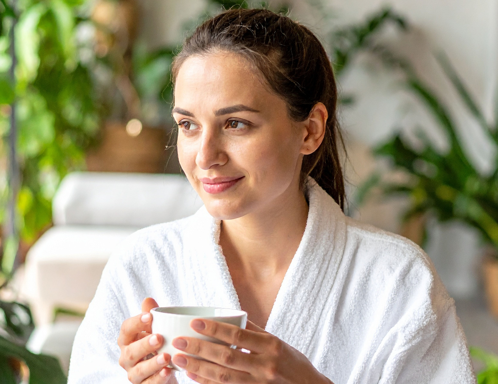 A woman with brown hair in a white robe holding a cup, sitting in a sunlit room with green plants in the background.