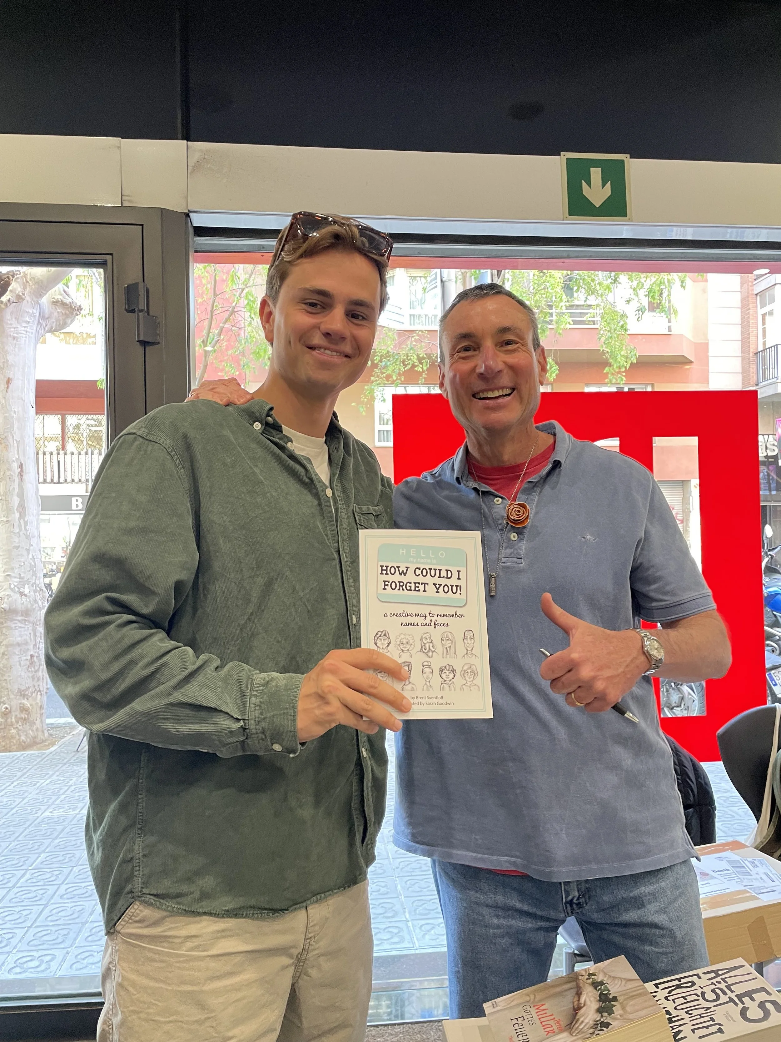 Two smiling men standing inside a retail store, one holding a book titled 'How Could I Forget You!' with illustrations of faces on the cover, and the other giving a thumbs-up gesture.