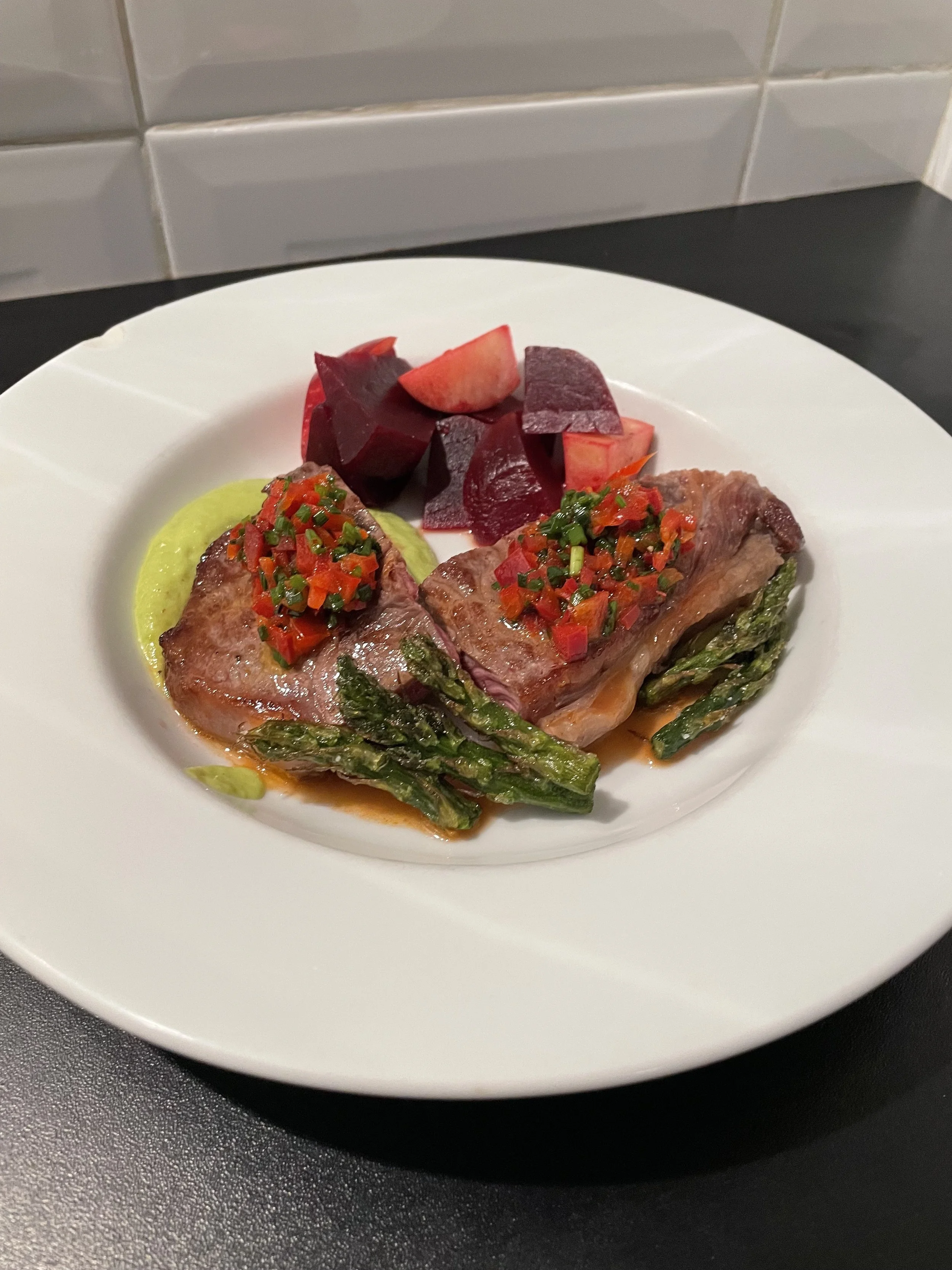 Plate with two cooked asparagus, two pieces of cooked meat topped with diced red pepper and chives, and chopped beets.