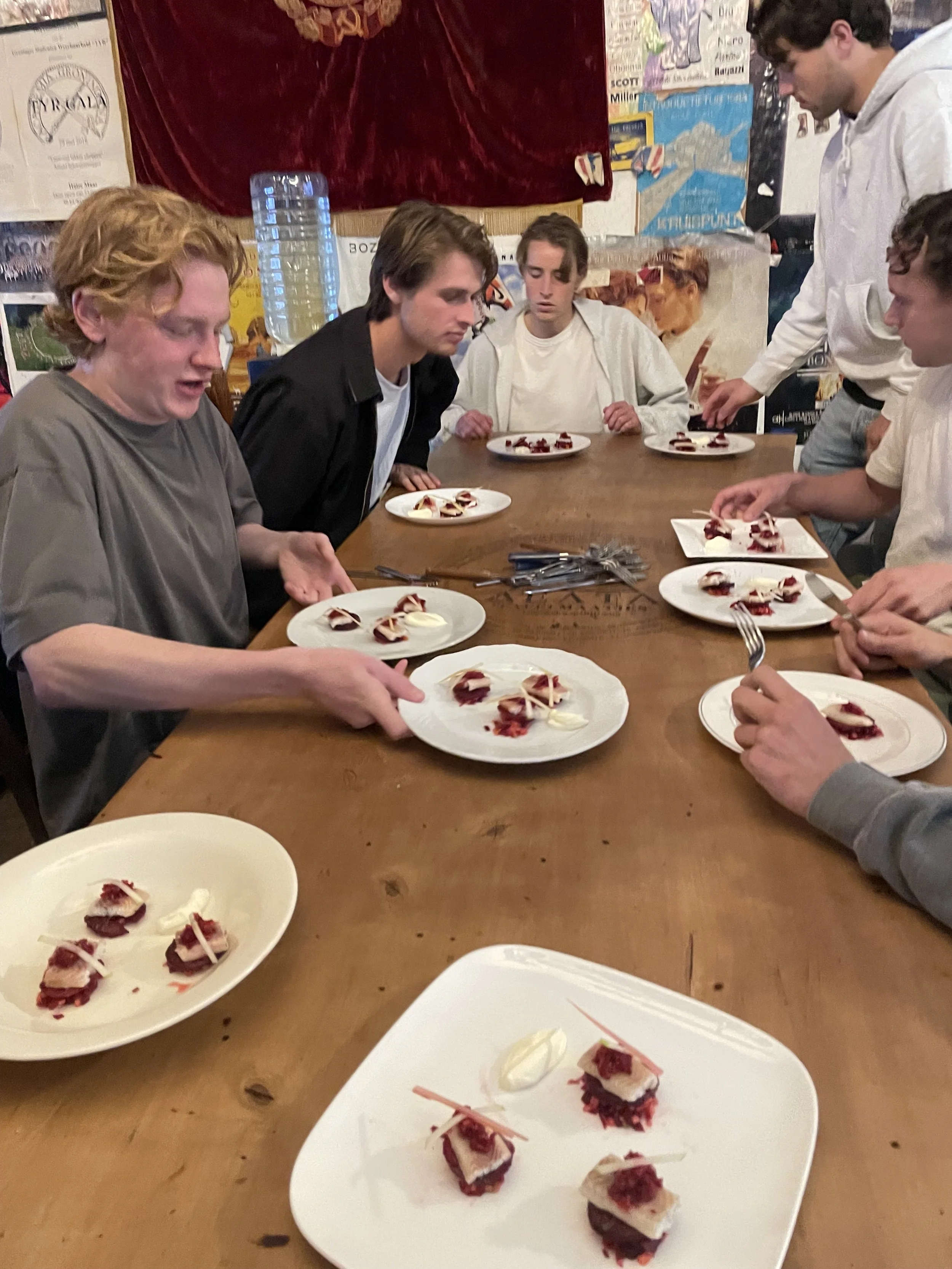 Group of people gathered around a wooden table, serving and eating small appetizers with various ingredients on white plates in a casual indoor setting.