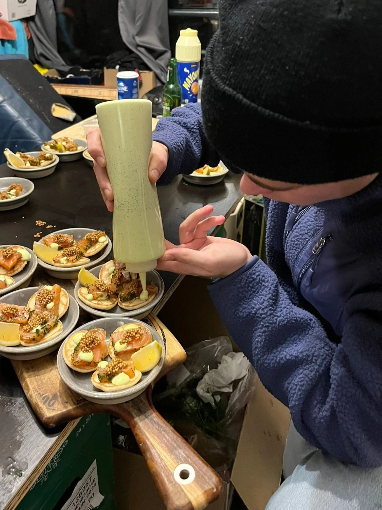 Person in a black beanie and blue jacket squeezing sauce onto small plates of open-faced sandwiches with smoked salmon, lemon wedges, and toppings on a wooden serving board.